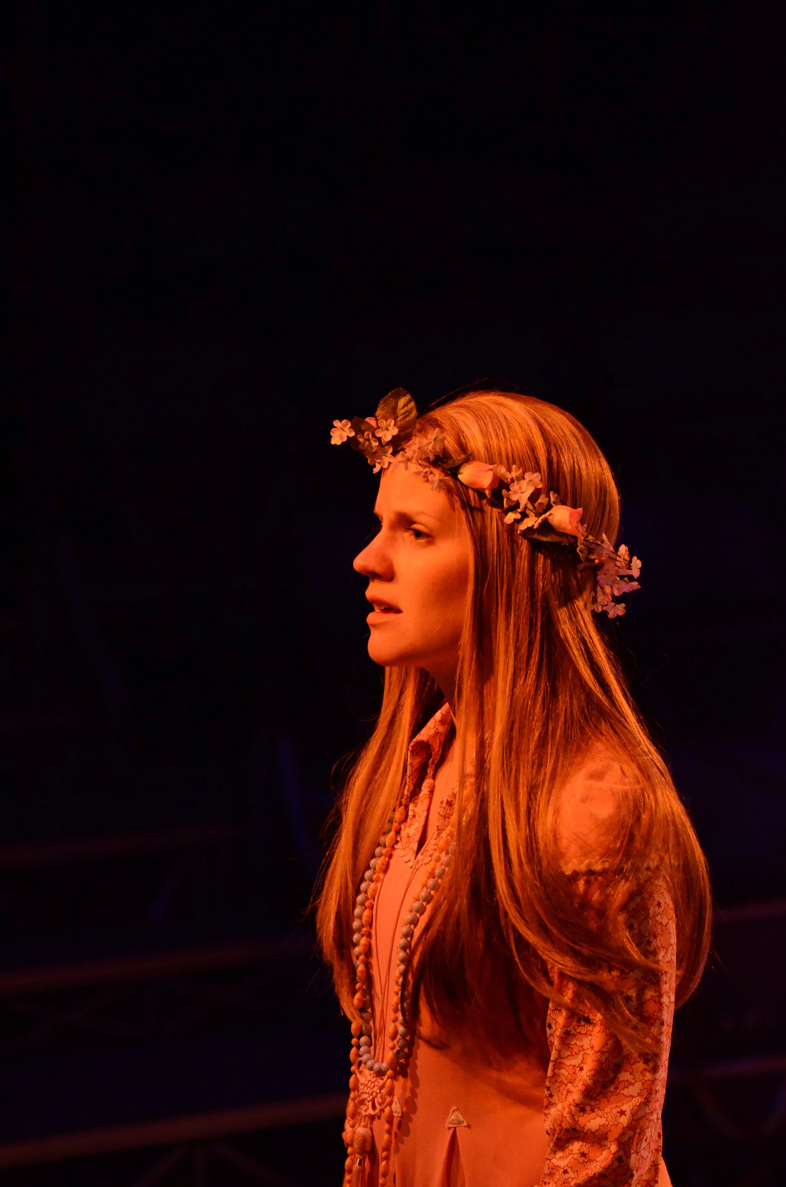 A woman with long red hair wearing a floral crown and traditional clothing, standing against a dark background.