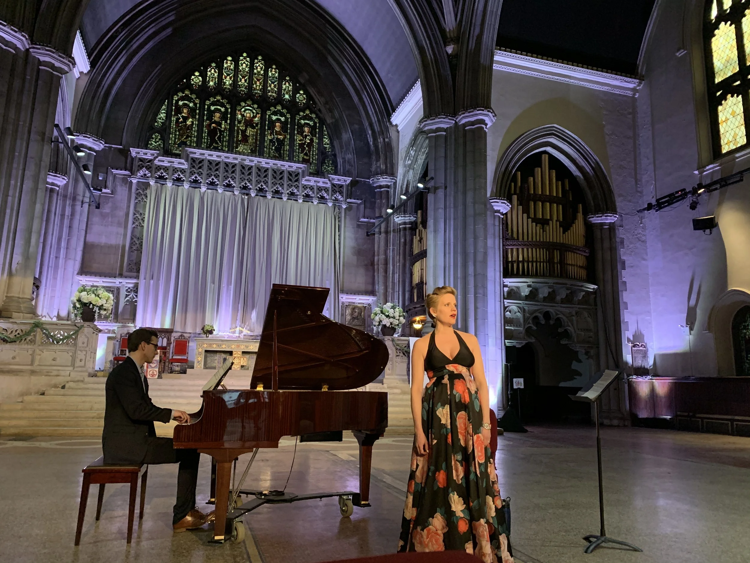 A woman singing at a microphone and a man playing a grand piano on a stage inside a church or cathedral with stained glass windows and Gothic architecture.