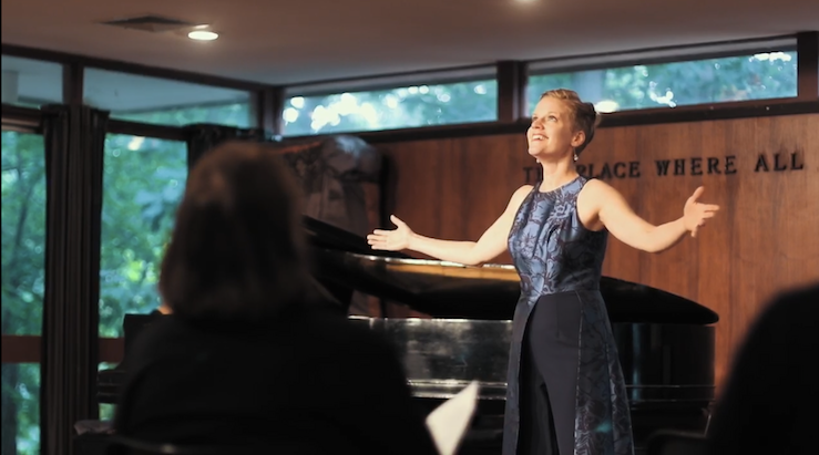 Woman in a blue dress performing or giving a speech in a room with large windows and a wooden wall.