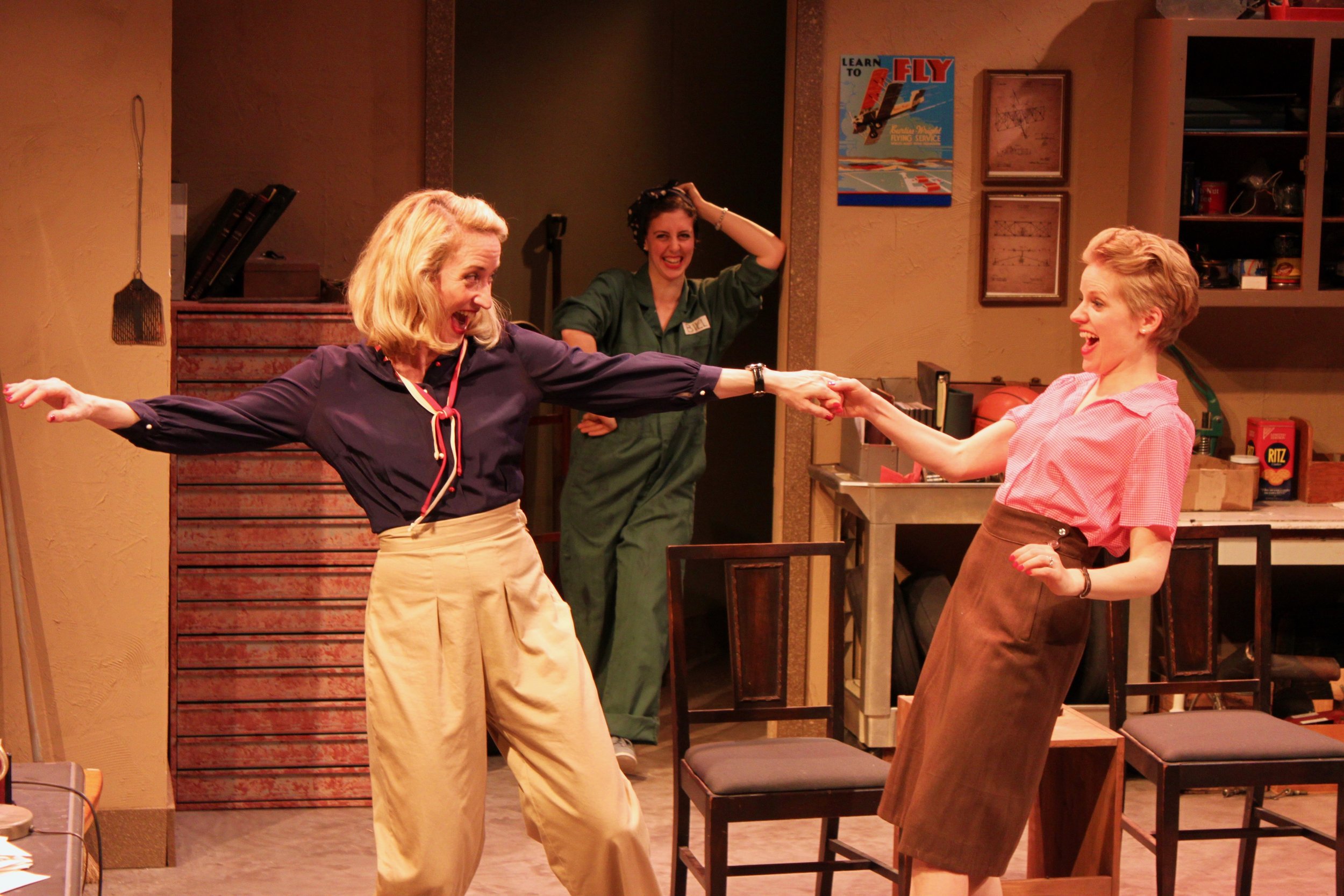 Two women are dancing happily and holding hands while another woman watches and smiles in a room with shelves and wall decorations.