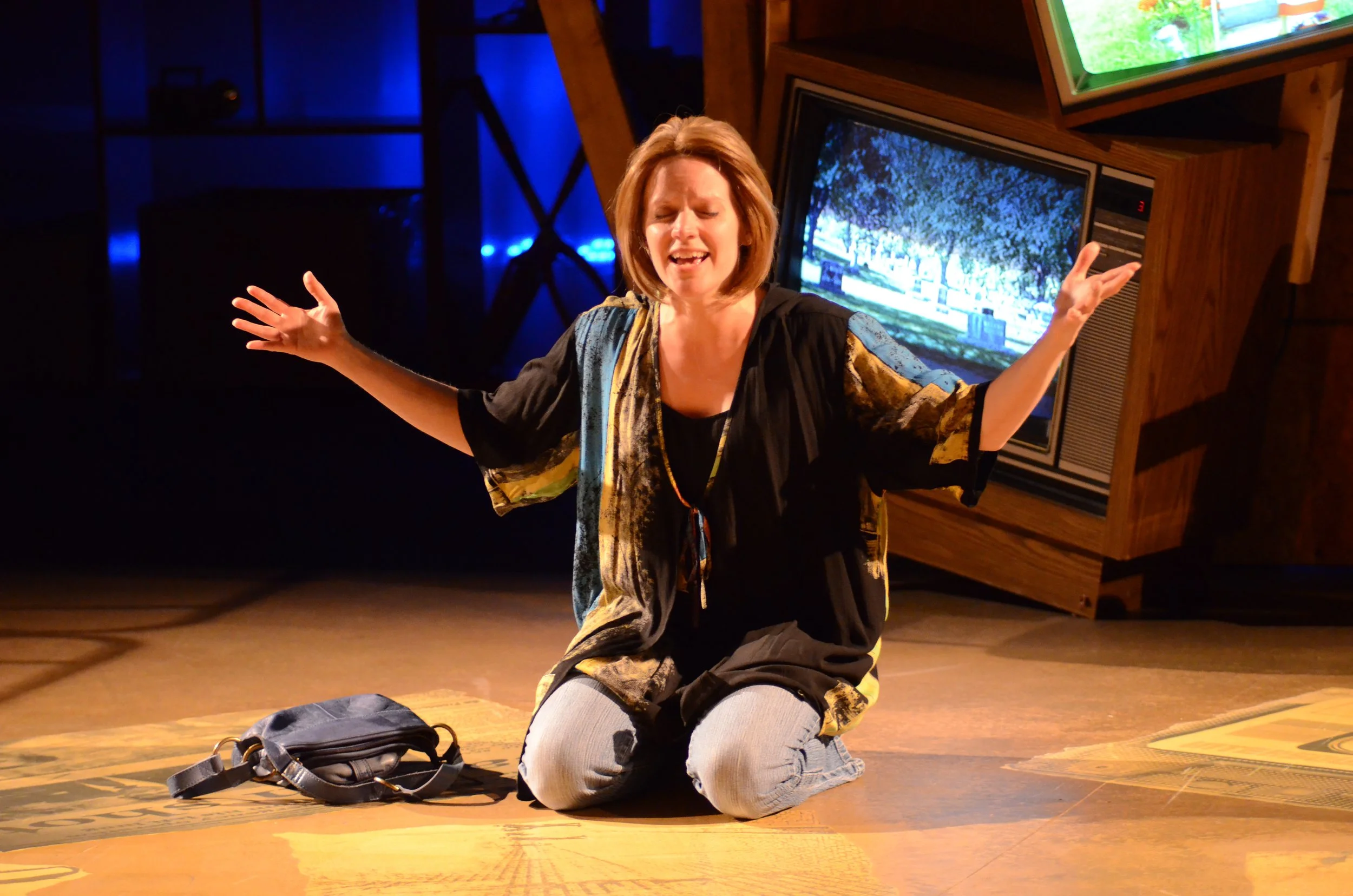 A woman kneeling on the floor with her arms spread wide, eyes closed, and a joyful expression. Behind her, there are television screens and dim lighting in a room that appears to be a theater or performance space.