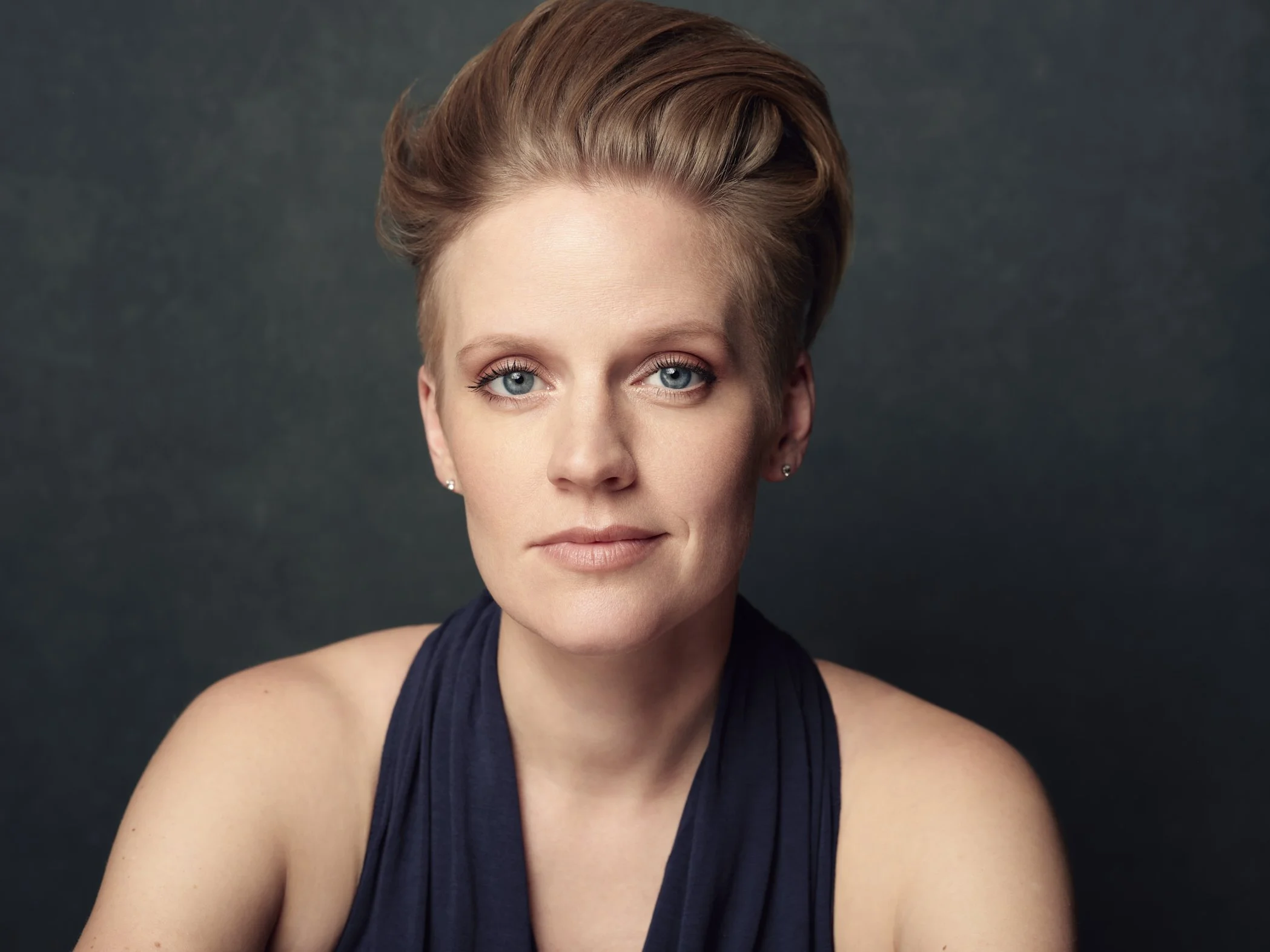 Close-up portrait of a woman with short, styled blonde hair, blue eyes, and light skin, wearing a sleeveless dark navy top against a dark textured background.