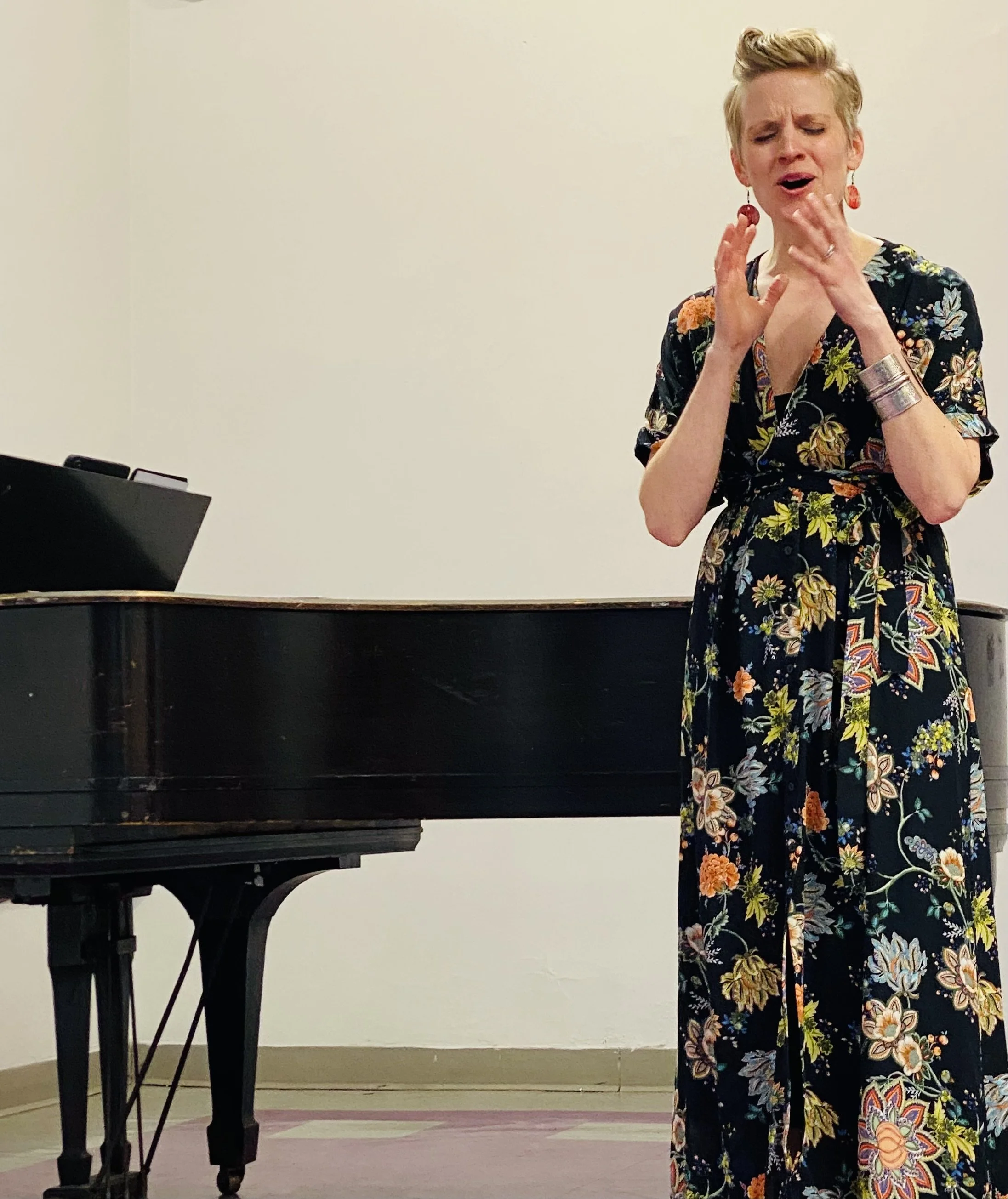 A woman singing passionately in front of a piano, wearing a floral dress and red earrings, in a room with a plain white wall.