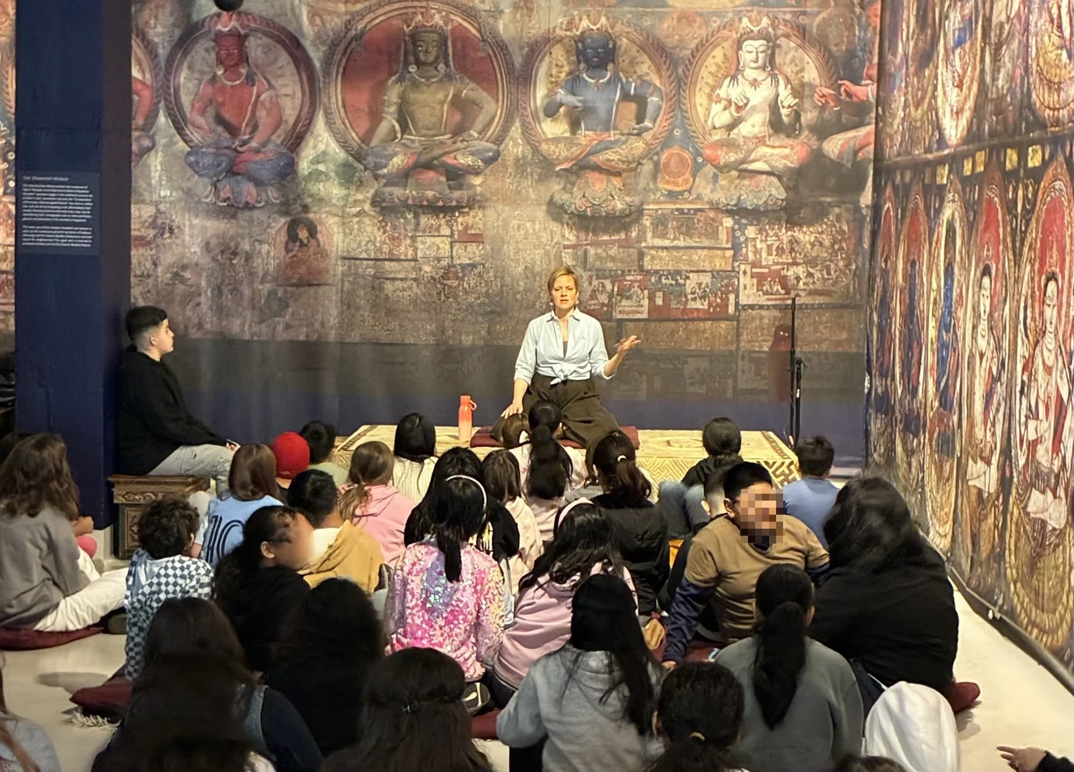 A woman sits on a cushion addressing a group of children seated on the floor in front of her, with a large mural of seated deities behind them.