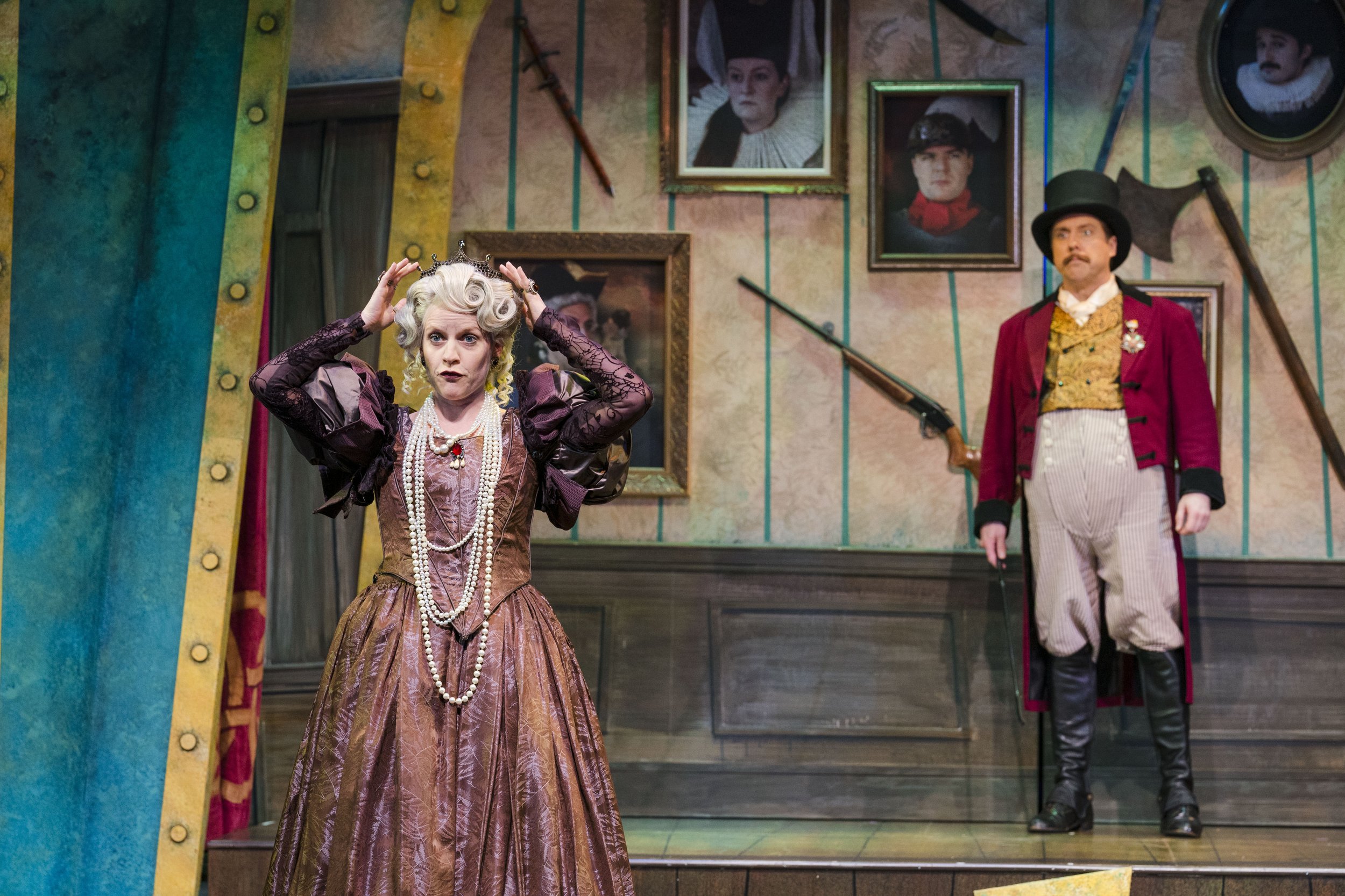 Actors in costume on stage, with the woman in the foreground wearing a brocade dress, pearls, and a tiara, and the man in the background wearing a top hat, red coat, and striped pants, surrounded by framed portraits and decorative swords.
