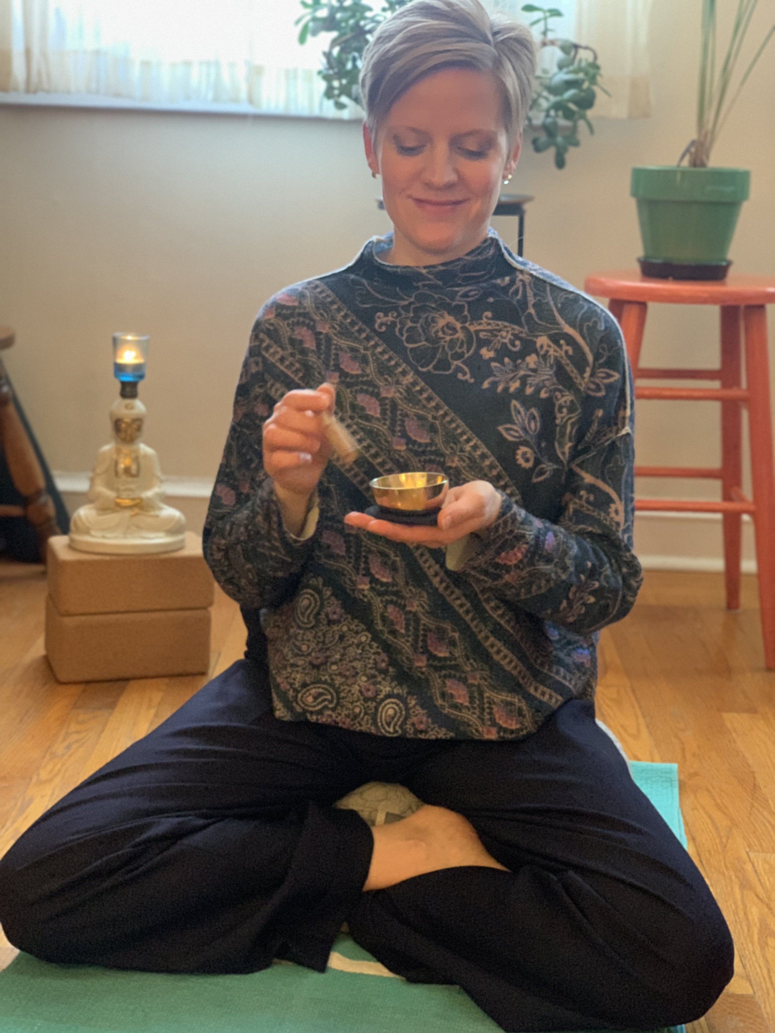 A woman with short blonde hair sitting cross-legged on a green yoga mat indoors, holding a meditation bell in her hands, with closed eyes and a peaceful expression.