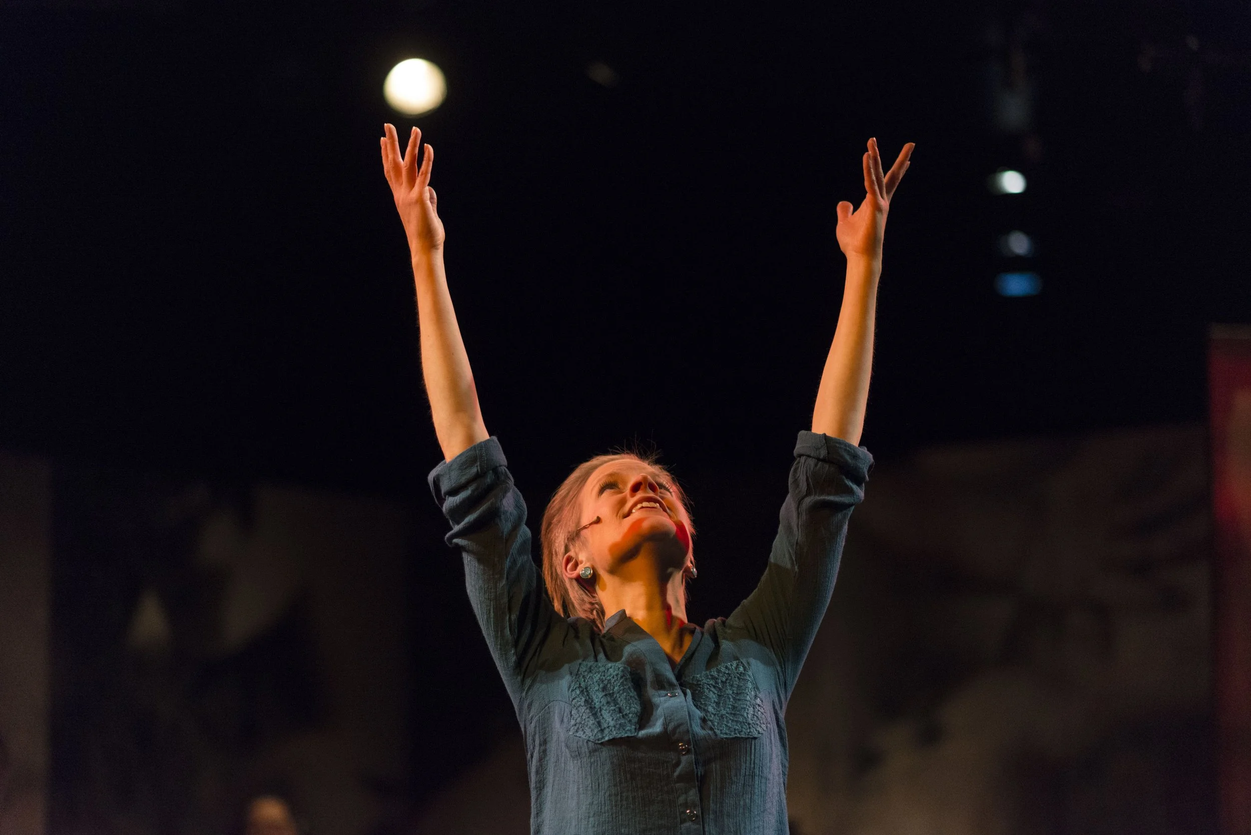 A woman raising her arms upward in a dark setting with a bright light visible in the background.