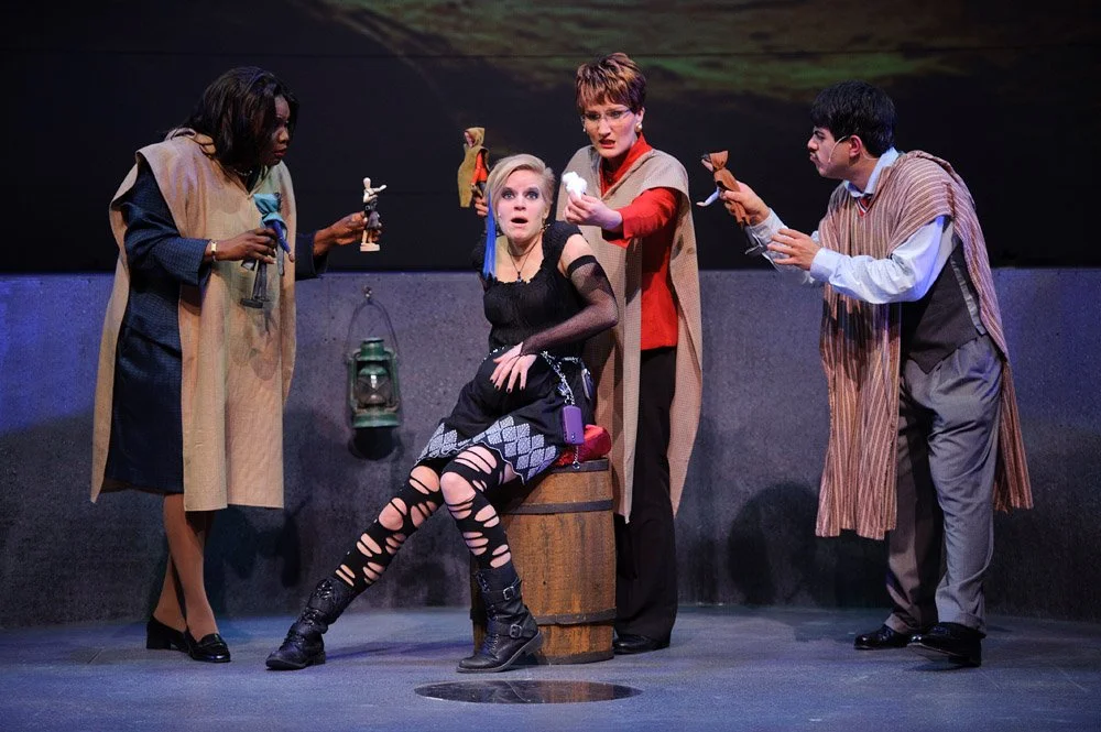 Stage scene with four actors performing. One woman seated on a barrel with a surprised expression, dressed in black torn clothes. Three other actors standing around her, holding puppets. The woman on the left holds a puppet resembling a dog or simila