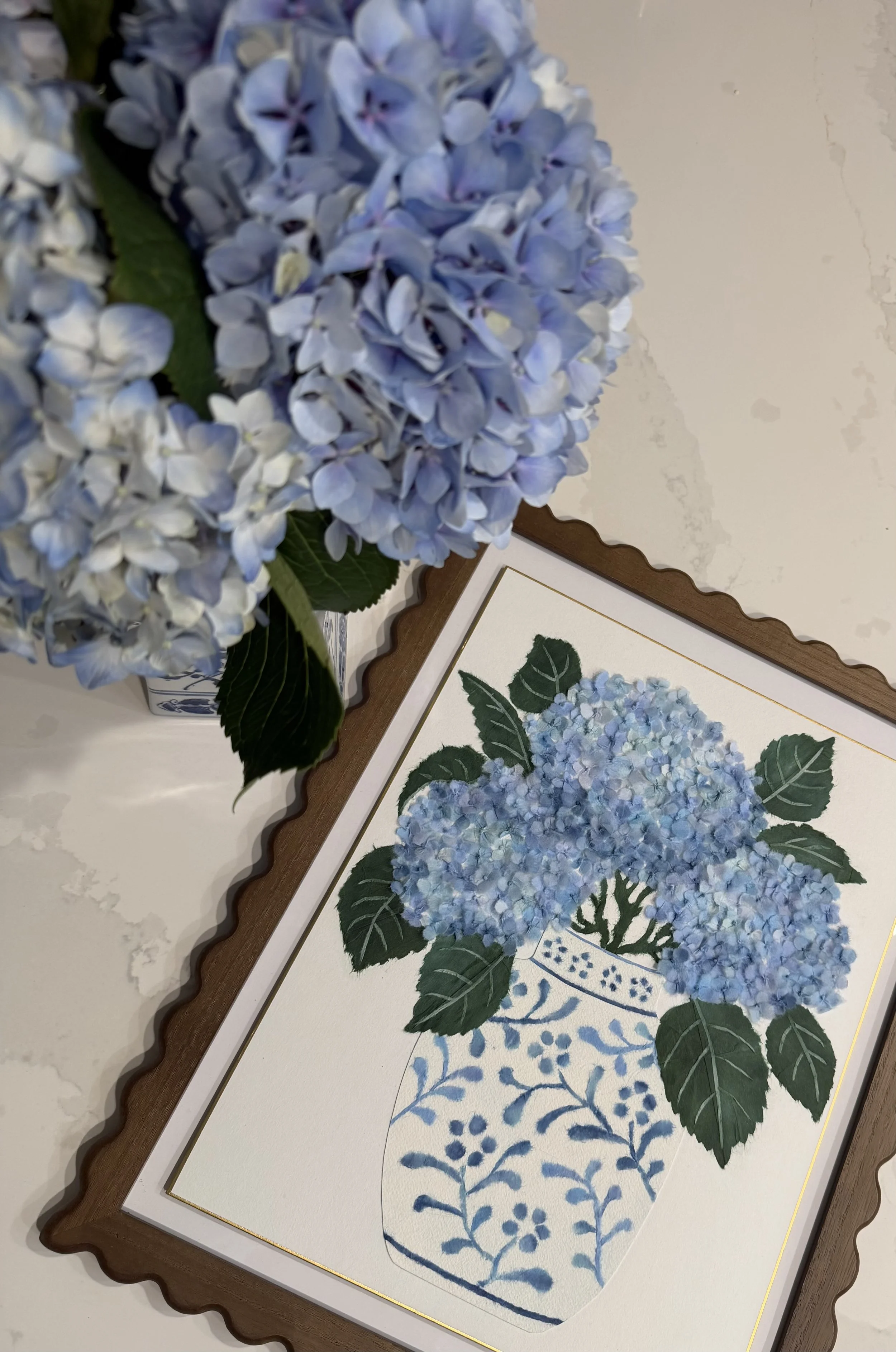 A bouquet of blue hydrangea flowers on a table and a framed botanical illustration of a hydrangea plant.