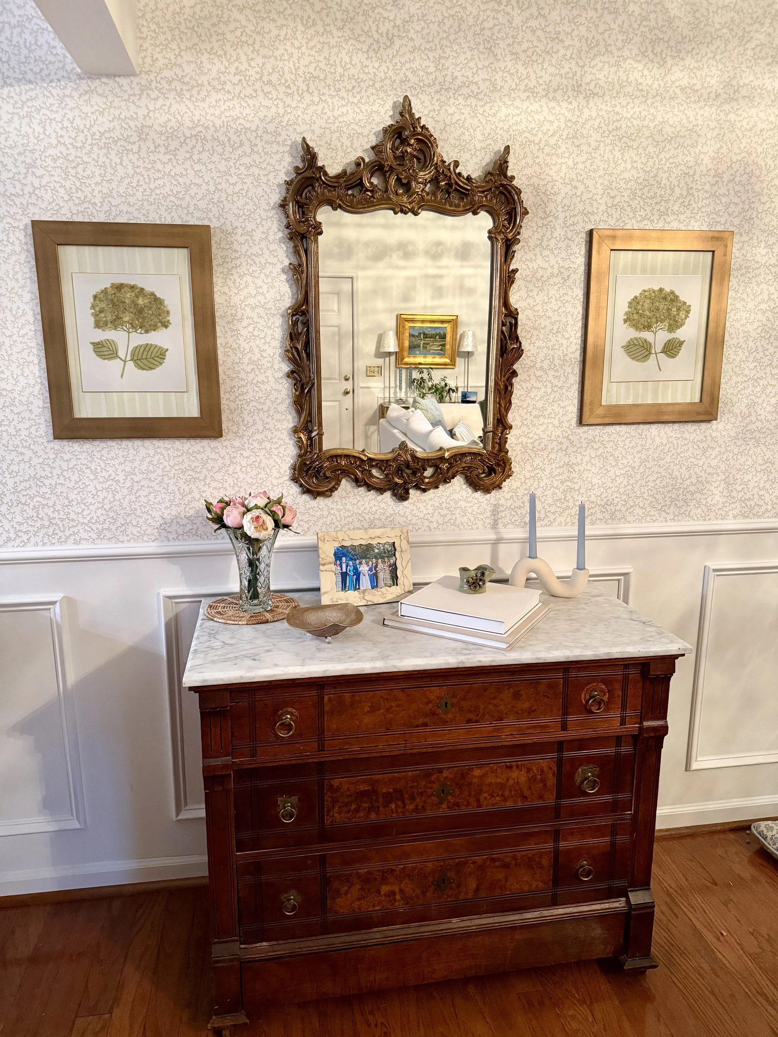 A wooden dresser with a marble top, with a vase of pink flowers, a framed family photo, books, and decorative objects on top, and a large ornate mirror hanging above. Matching green hydrangea artwork is on either side.