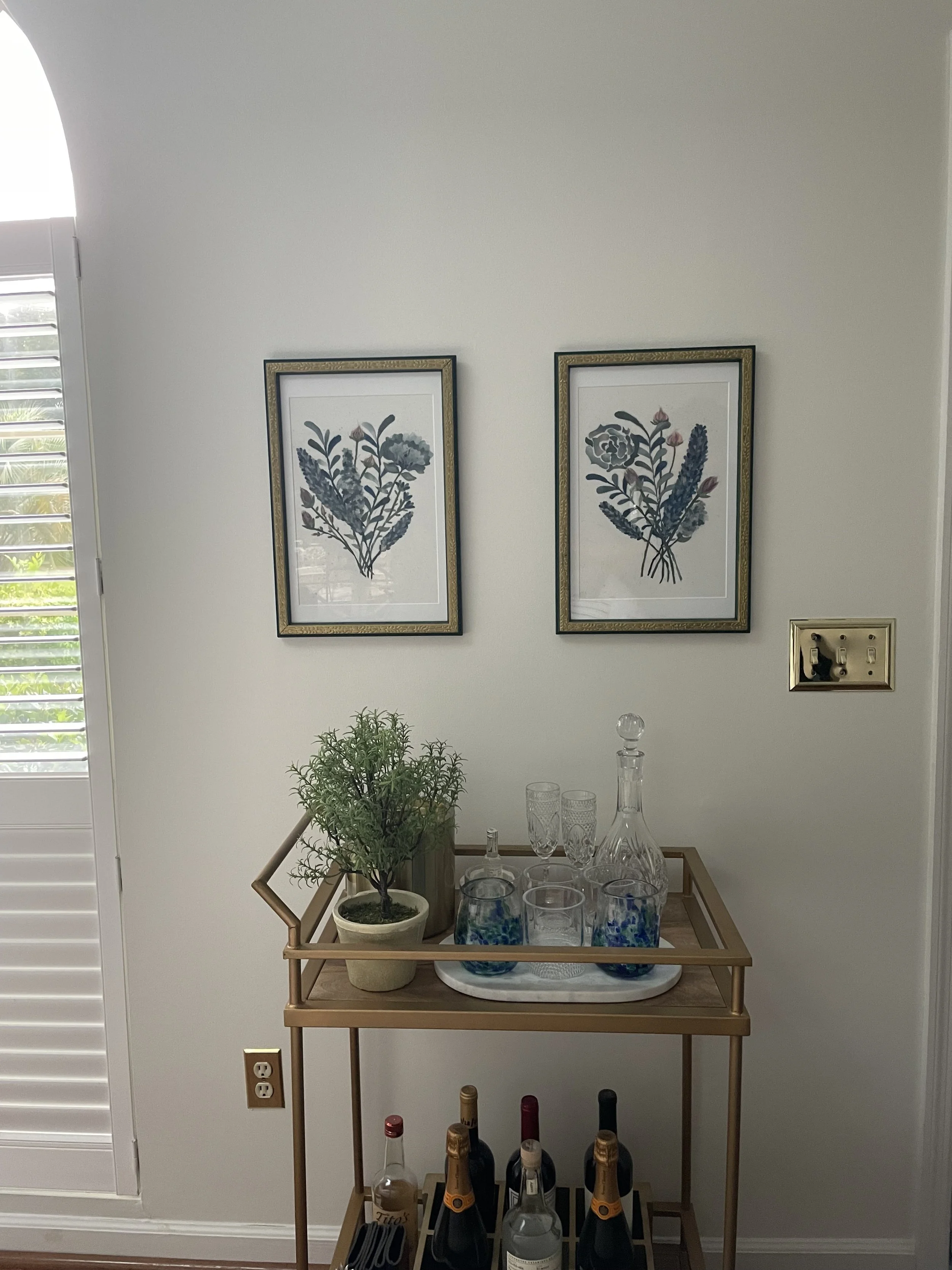 A bar cart with bottles, glassware, and decorations, placed against a white wall with two framed floral art prints, near a window with white plantation shutters.