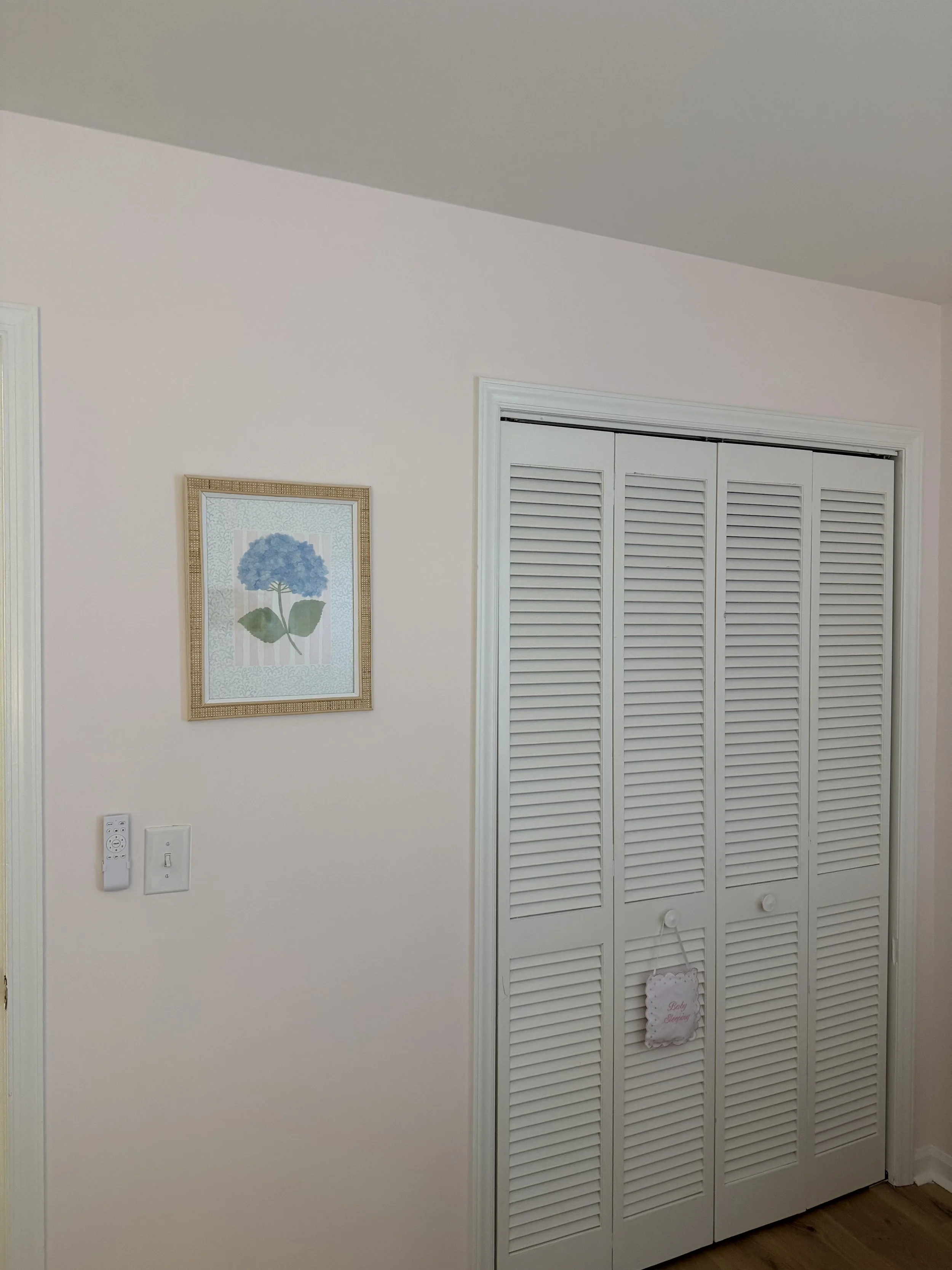 White louvered closet doors in a corner of a room with a pink wall, framed artwork of a blue hydrangea flower, and a piece of decorative paper with 'Baby Sleeping' text hanging on the door.