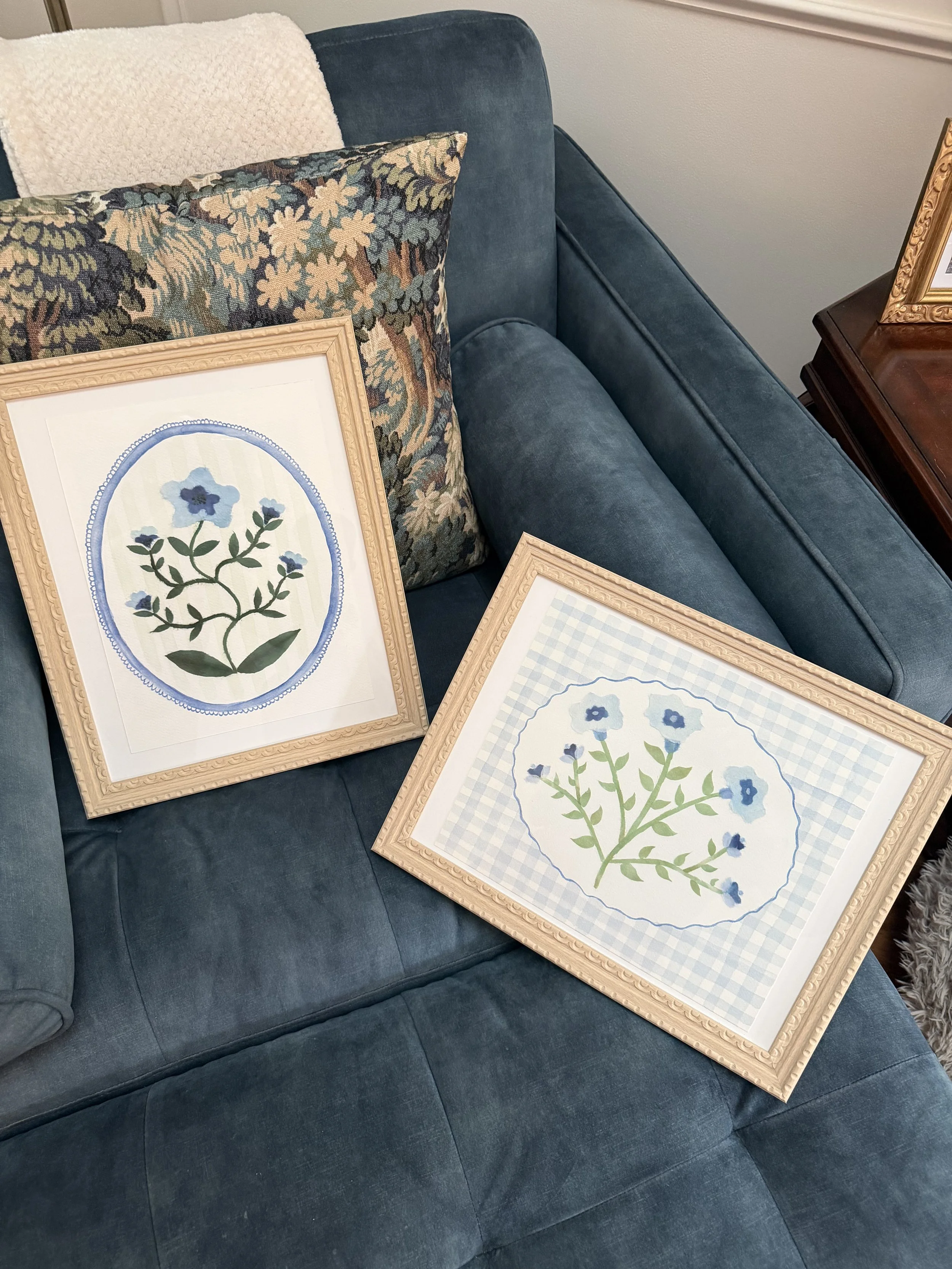 A blue velvet sofa with three framed botanical prints with floral artwork resting on it, and a white textured pillow is visible. Part of a wooden side table with a decorative framed picture is to the right.