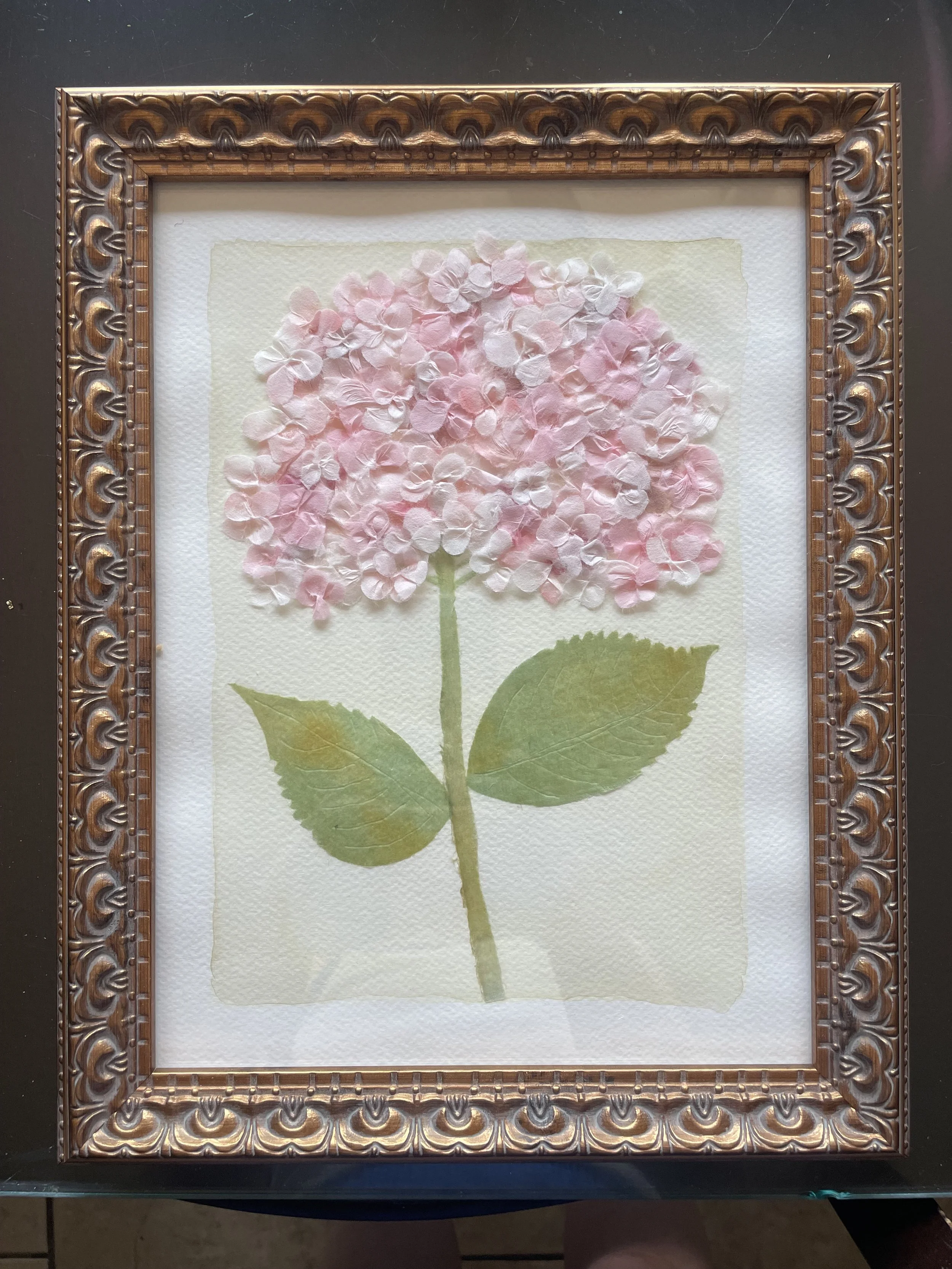 Artwork of a pink hydrangea flower with green leaves, framed in an ornate gold-colored frame, on a dark surface.