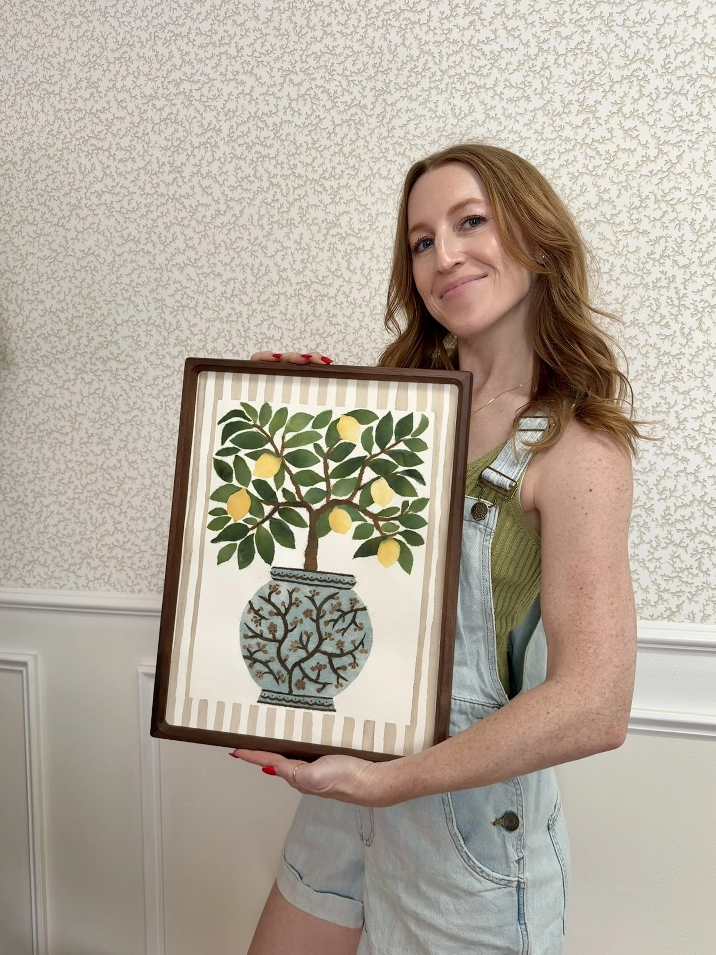 A woman with red hair, wearing a green top and overalls, holding a framed botanical illustration of a lemon tree with green leaves and yellow lemons, against a background with patterned wallpaper.