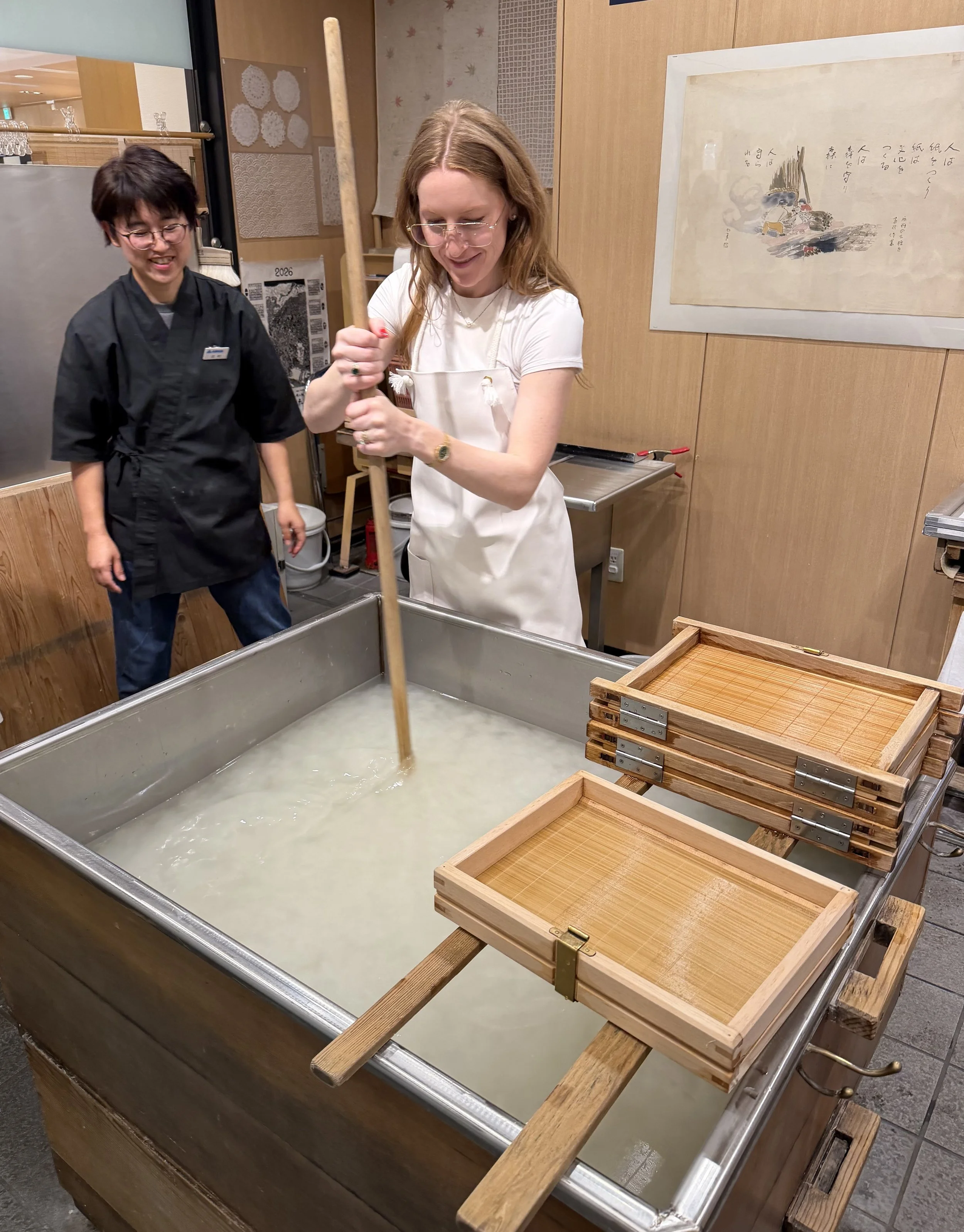 A woman with red hair in a white apron using a wooden tool to make washi paper in a large metal tub, with a young Japanese woman with short black hair in a black uniform watching and smiling.