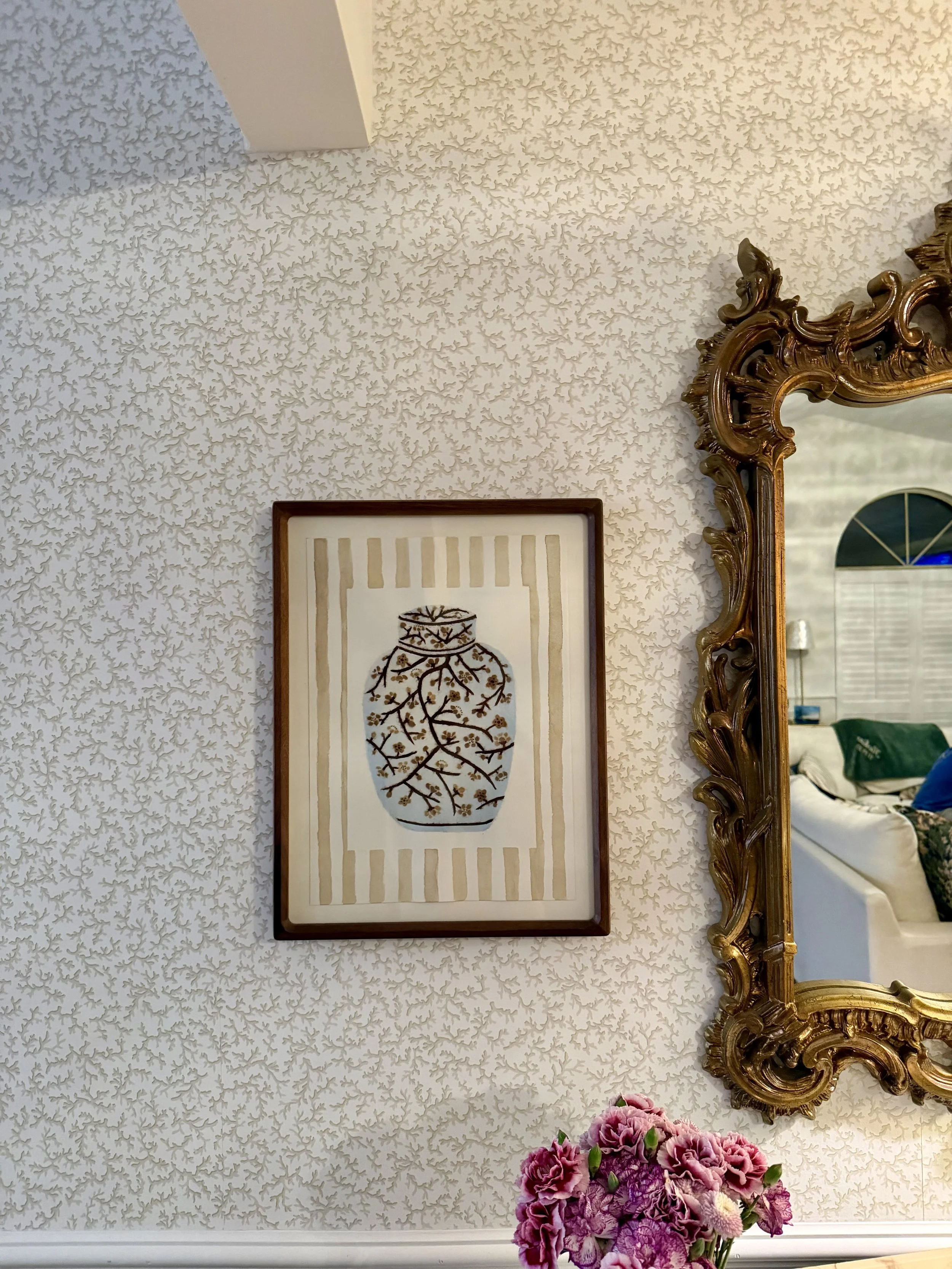 Decorative living room wall with ornate gold mirror, framed artwork of a ginger jar vase with branches and flowers.
