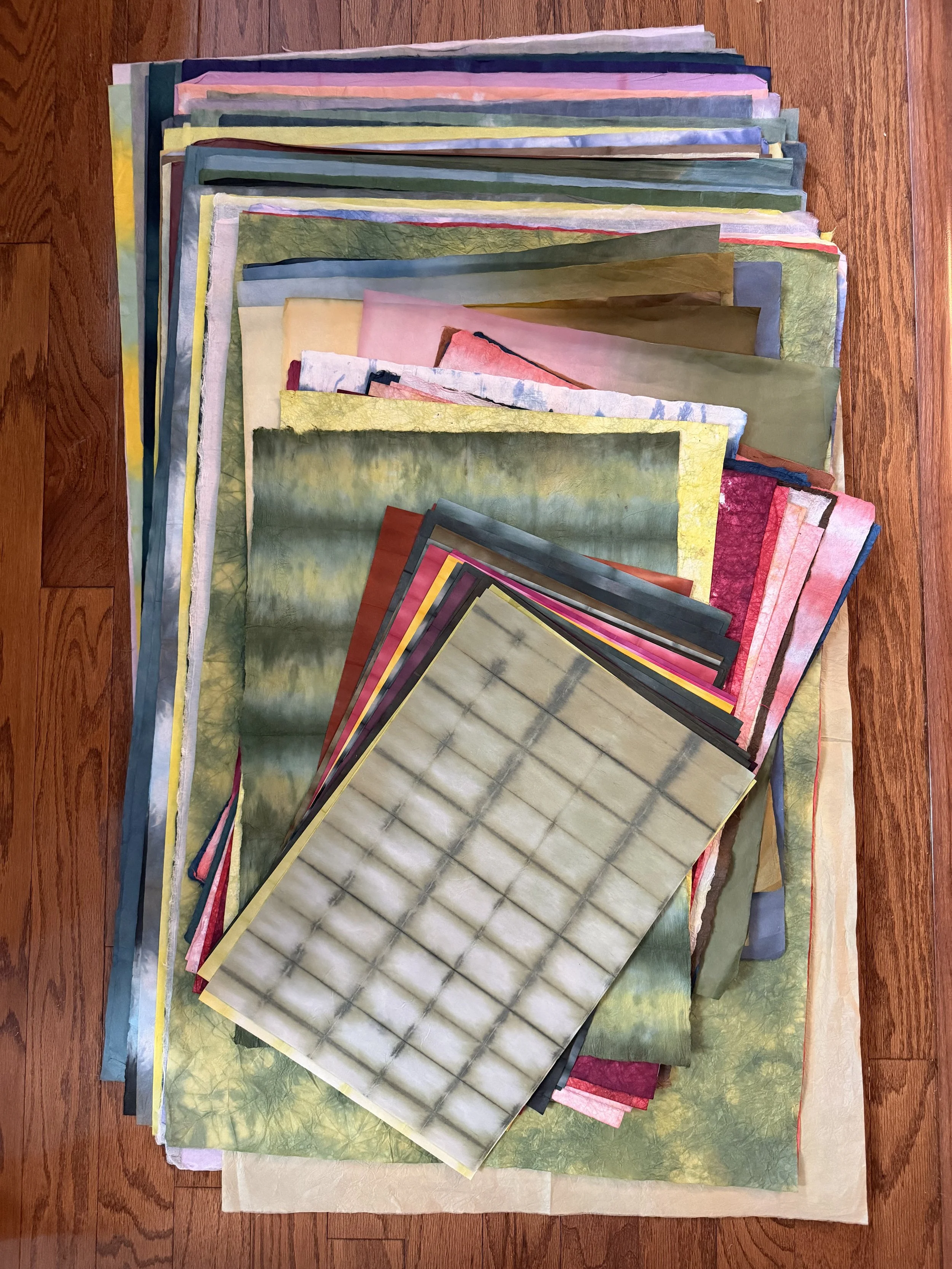 A collection of colorful washi paper sheets spread out on a wooden surface, arranged in overlapping layers.