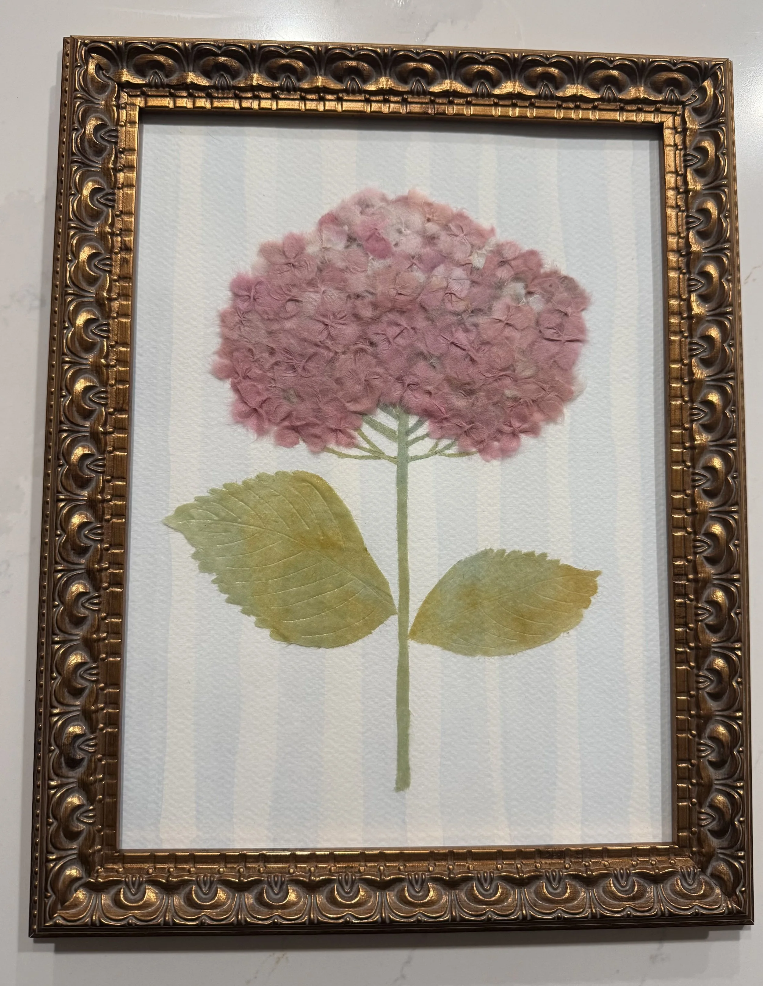 Framed artwork of a hydrangea flower with pink and purple petals with green leaves and stem.