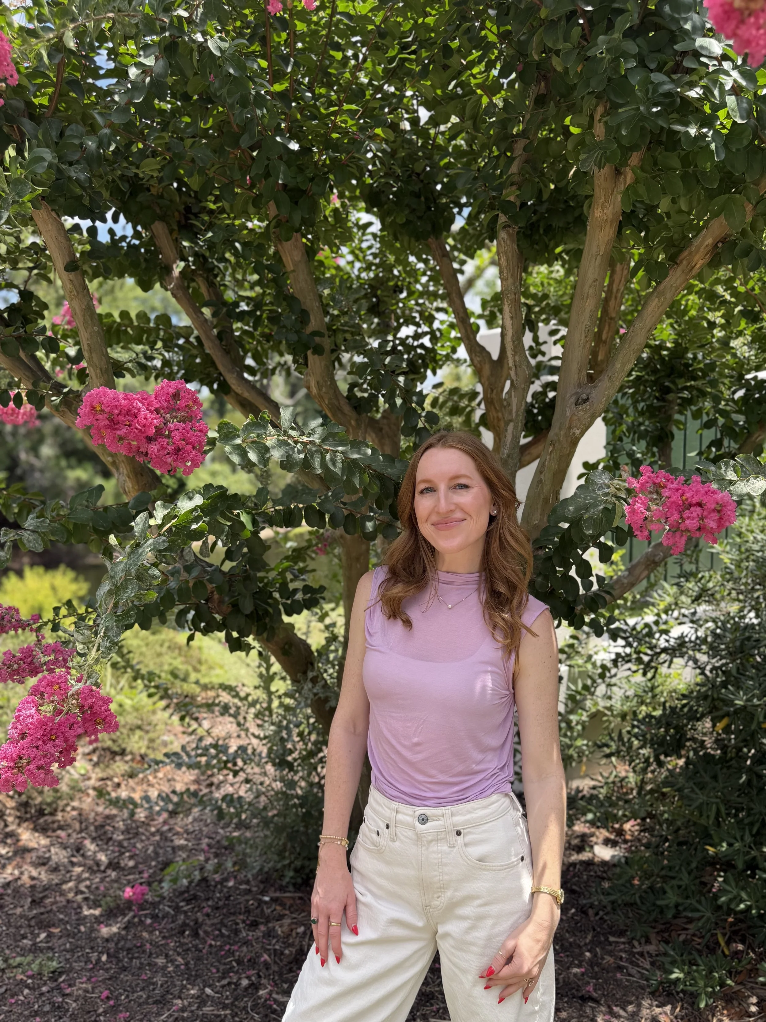 A woman with long wavy reddish-brown hair standing outdoors in front of a lush green tree with pink flowers. She is wearing a sleeveless lavender top and white pants, smiling softly at the camera.