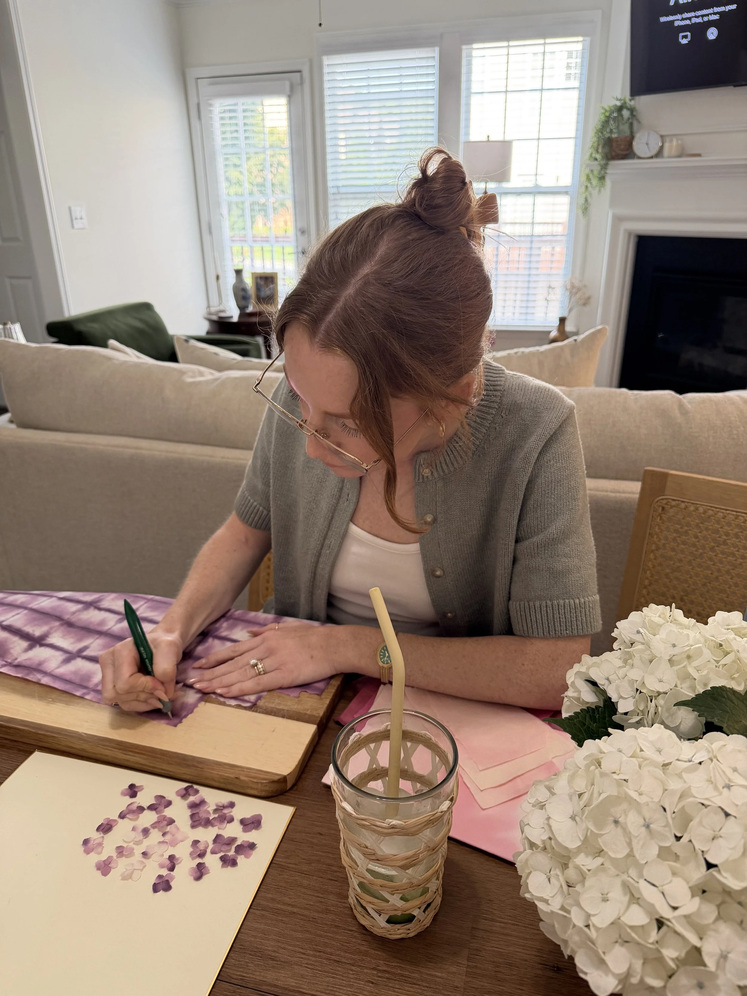A woman with red hair in a bun and wearing glasses is at a table, drawing on purple and pink paper. The table has hand torn flower petals on paper and a bouquet of white hydrangeas. The background shows a living room with large windows, a fireplace