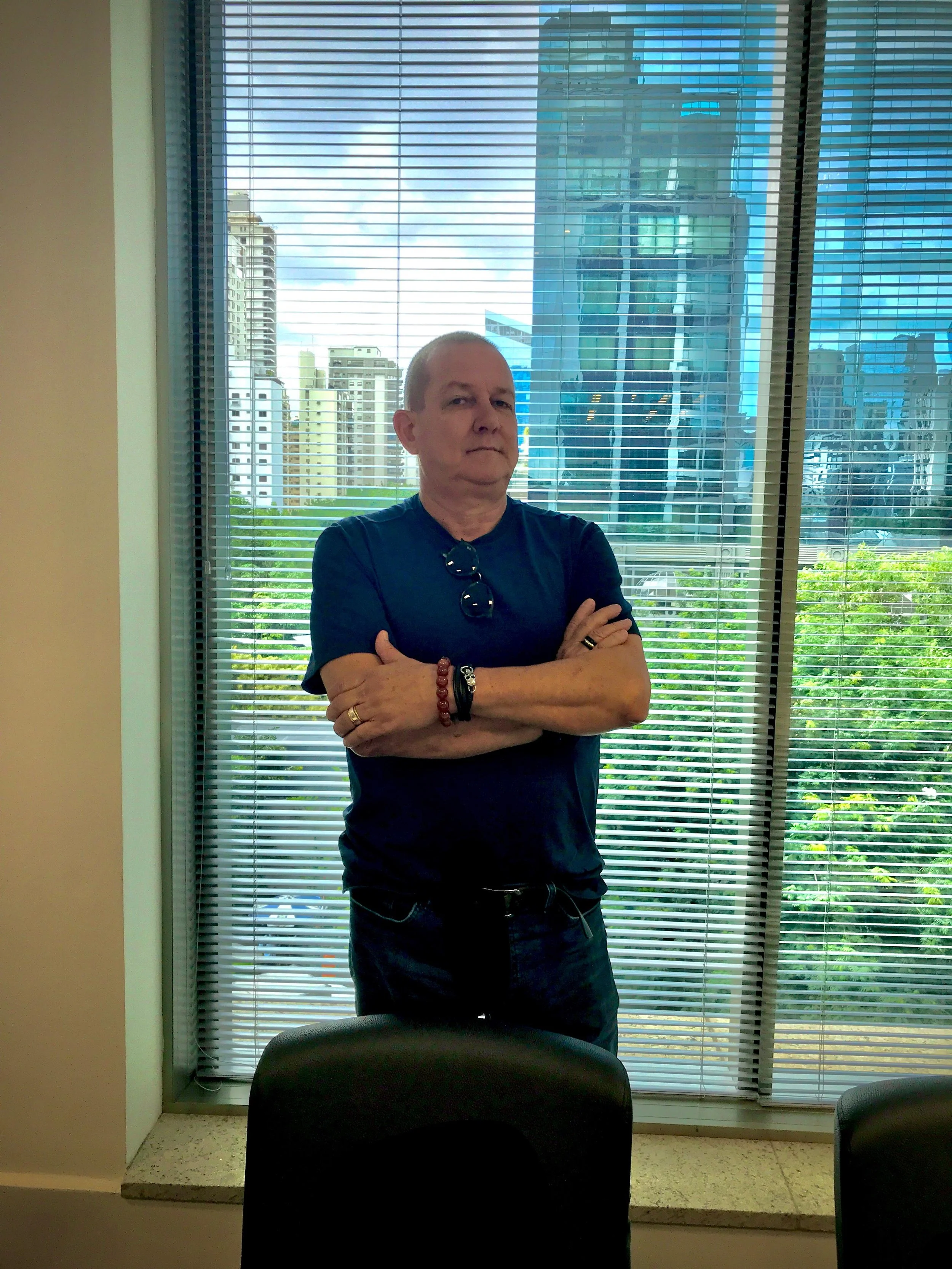 Homem com braços cruzados usando camiseta preta e óculos pendurados no pescoço, posando próximo a uma janela com cortinas de persianas horizontais, no interior de um edifício, com uma cidade de prédios visível ao fundo.