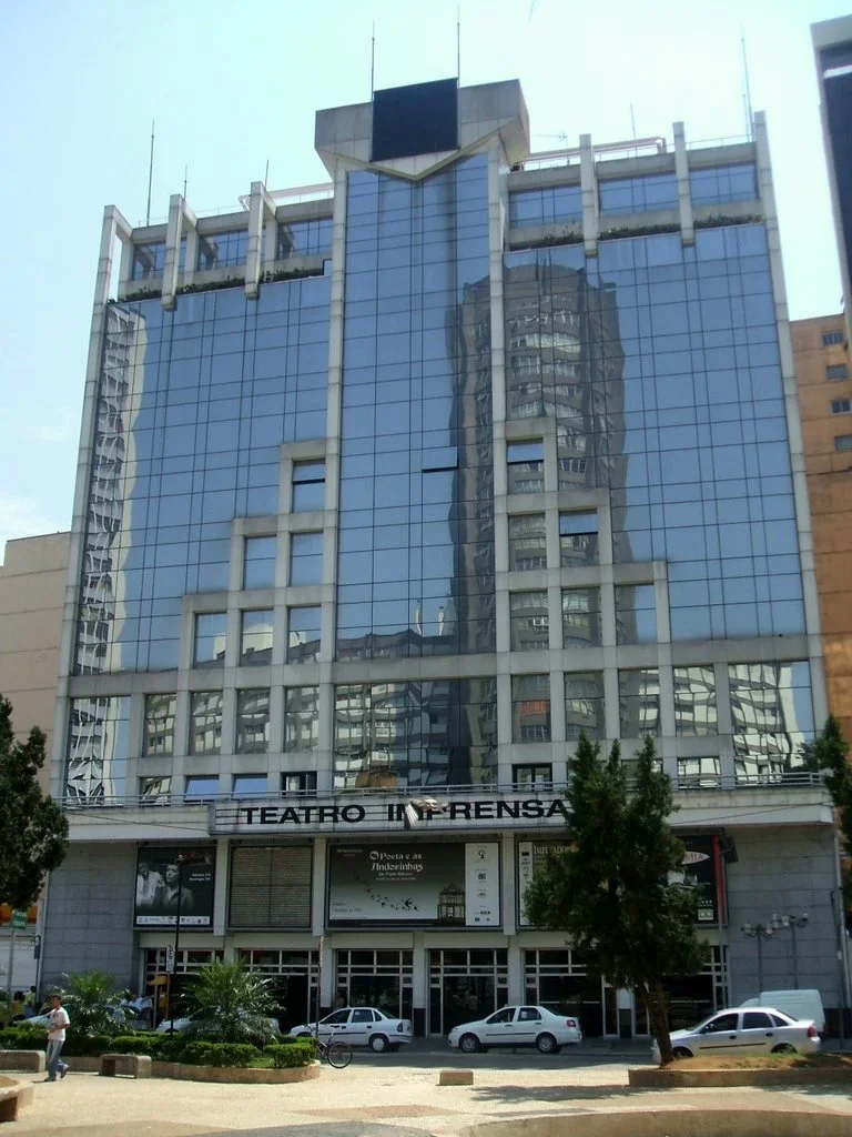 Prédio de alta altura com fachada de vidro que reflete outros edifícios da cidade, com uma assinatura na frente que diz 'TEATRO IN PENS'.