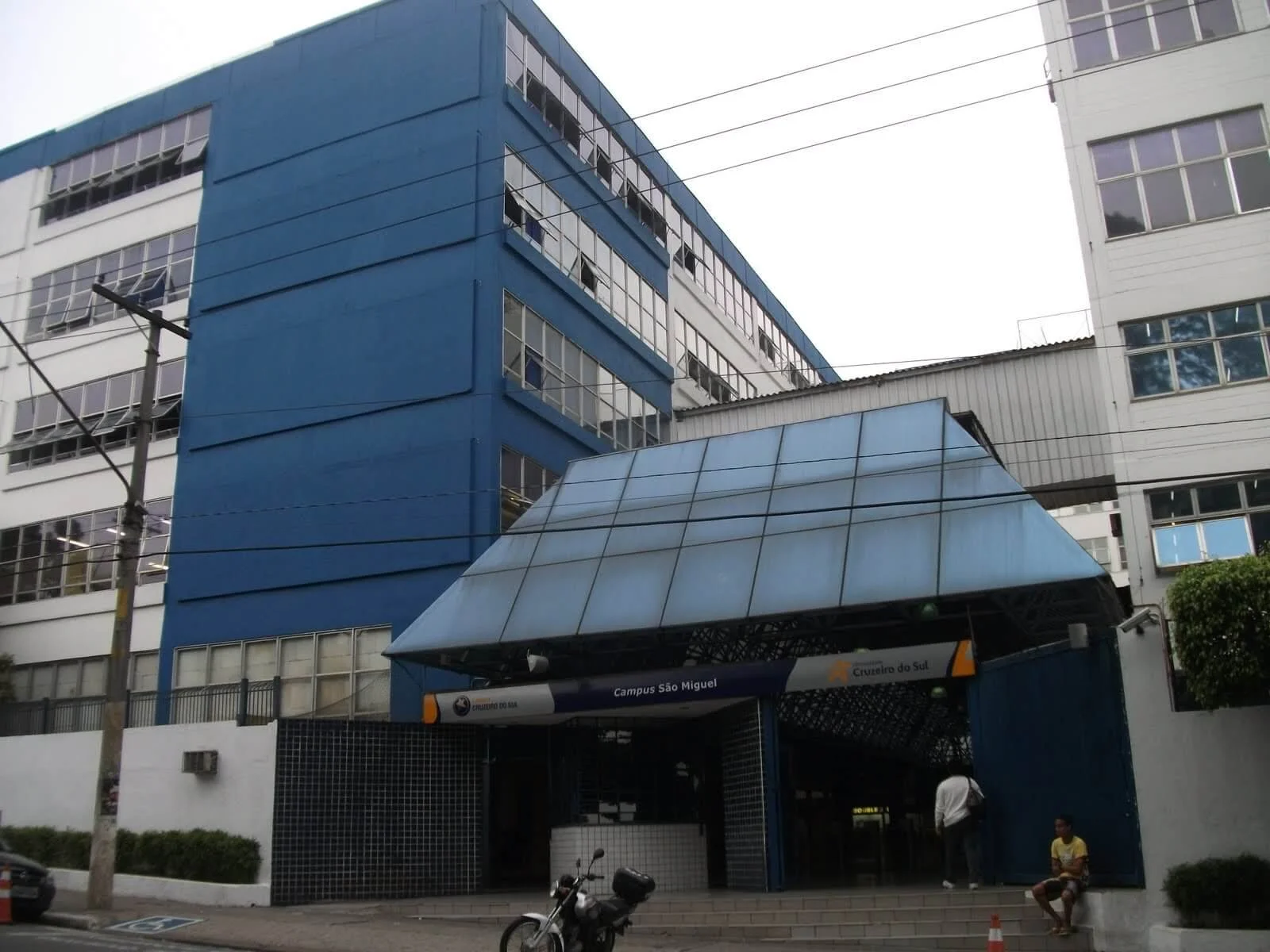 Entrada do campus São Miguel do Cruzeiro do Sul, com edifícios altos azuis e brancos, escadas na frente, e pessoas próximas à entrada.
