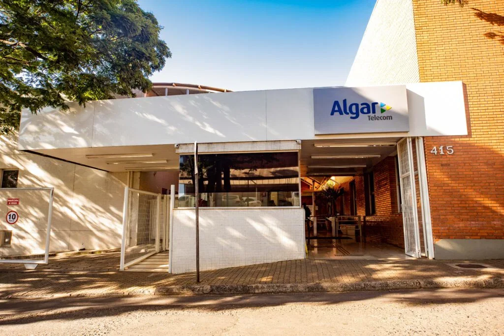 Edifício residencial com fachada de tijolos, entrada com acesso por portão de metal, e sinalização da Algar Telecom na parede da frente.
