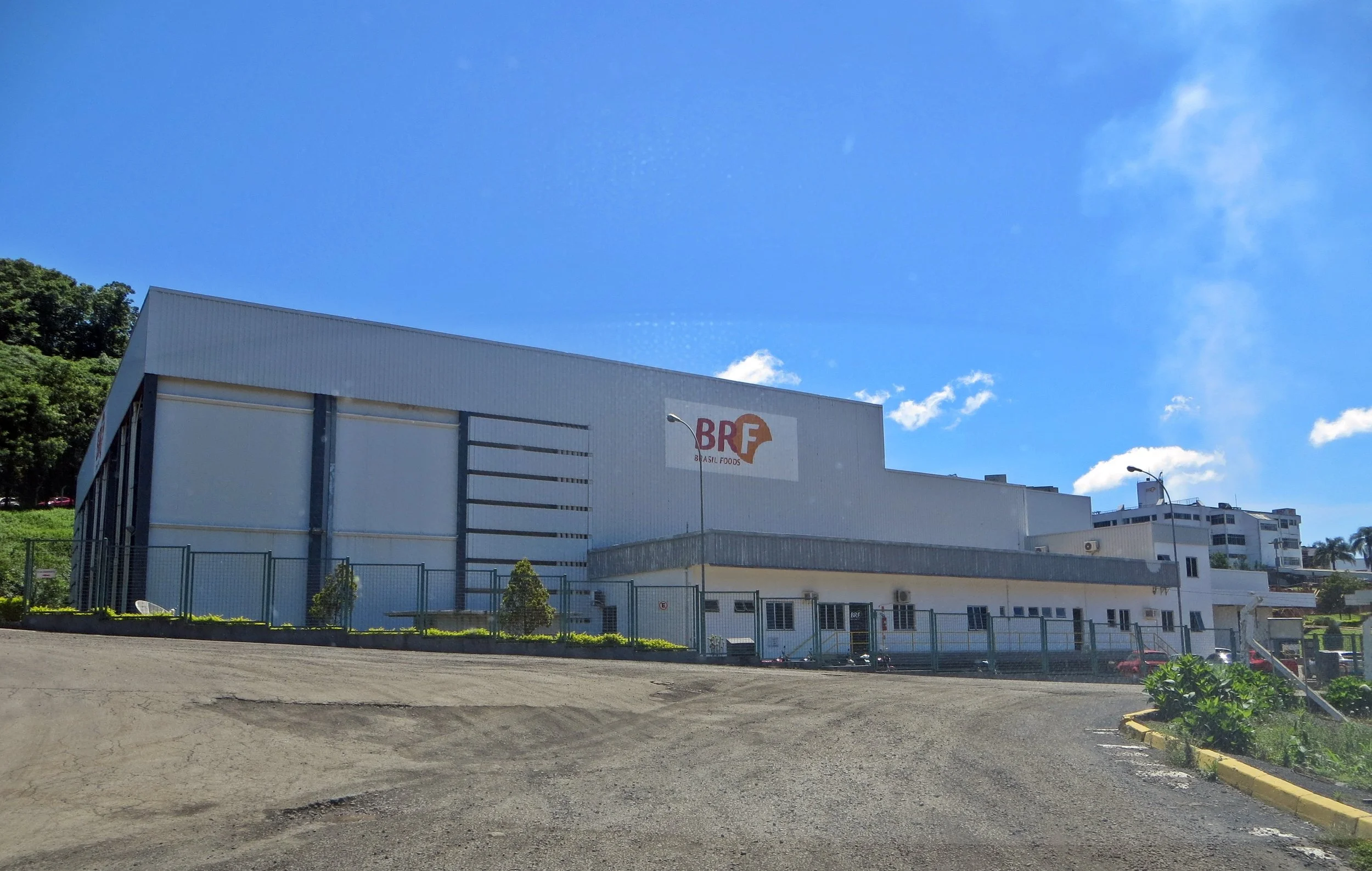 Fábrica com logo BRF (Brasil Foods) localizada em um terreno com cerca de metal, próximo a uma rua de piso não asfaltado e edifícios residenciais ao fundo. Céu claro com algumas nuvens.