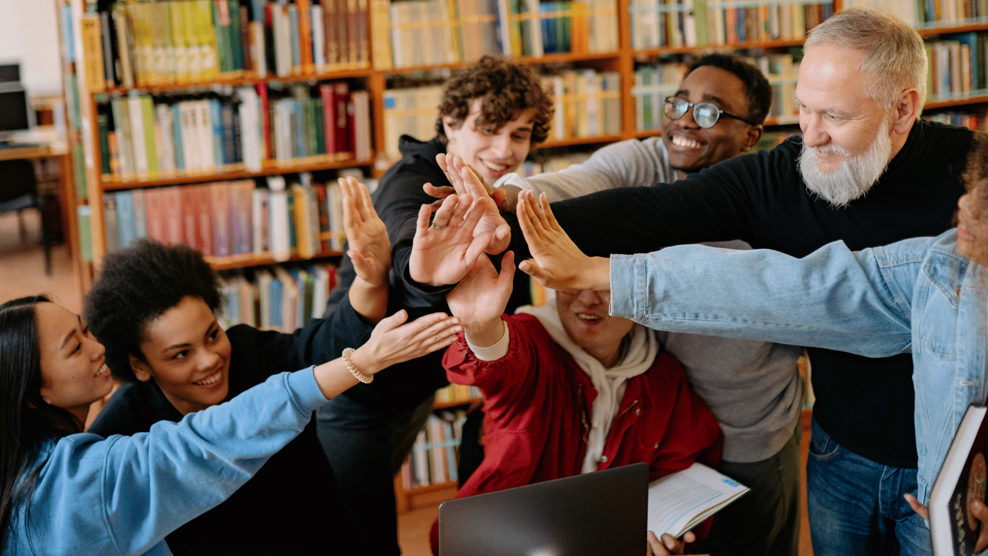 Diverse group of students smiling and bringing their hands together in a team huddle while studying in a library.