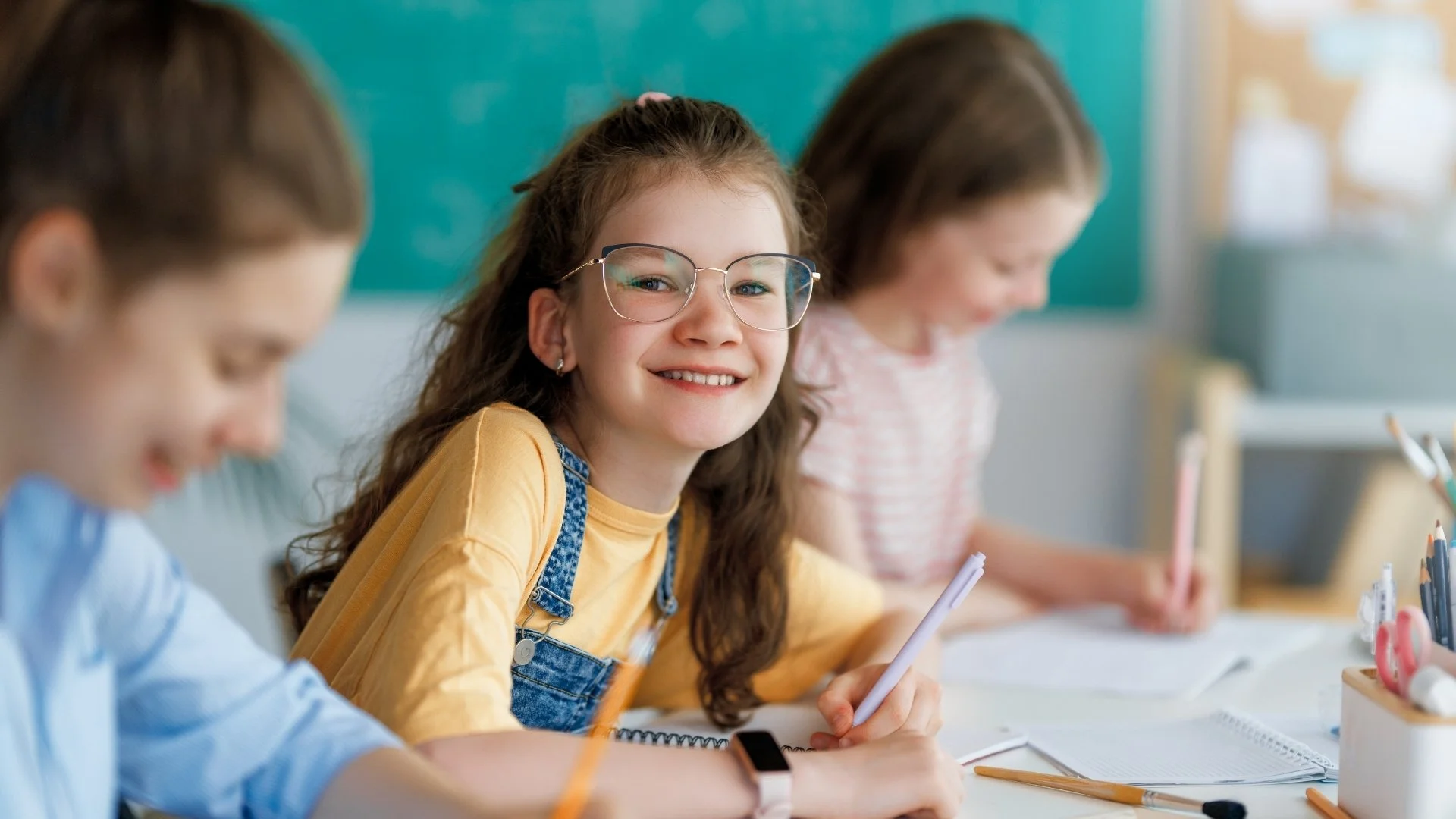Smiling elementary school student wearing glasses and writing in a notebook alongside classmates in a bright classroom.