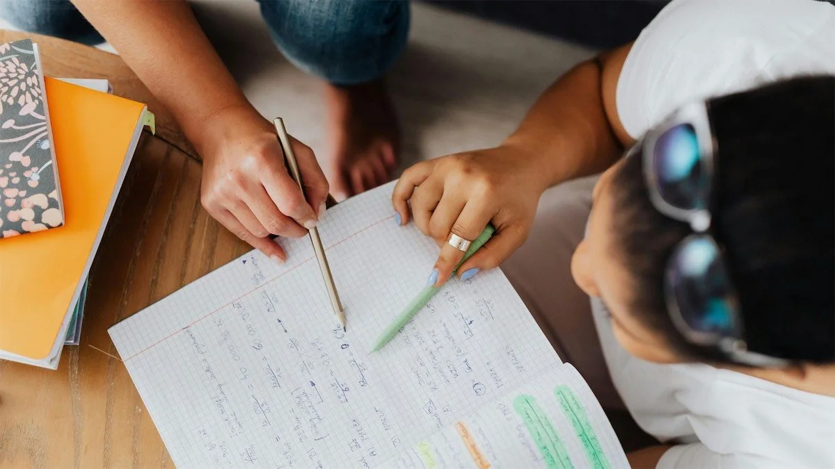 Two people working on homework or notes together at a wooden table, with notebooks and pens, highlighting and pointing at handwritten notes.