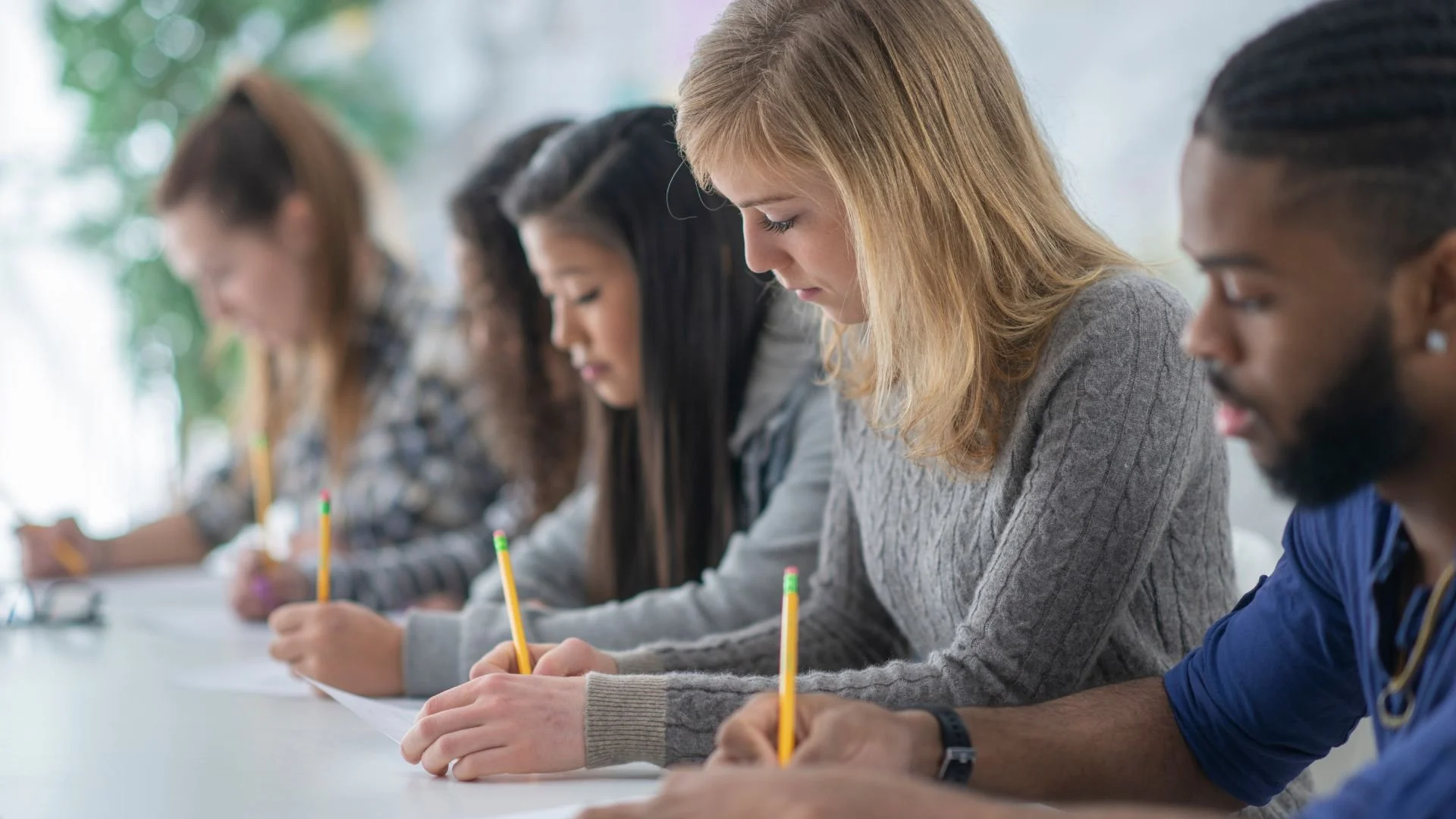 A group of young adults sitting at a table, writing with yellow pencils in a classroom or workshop setting.