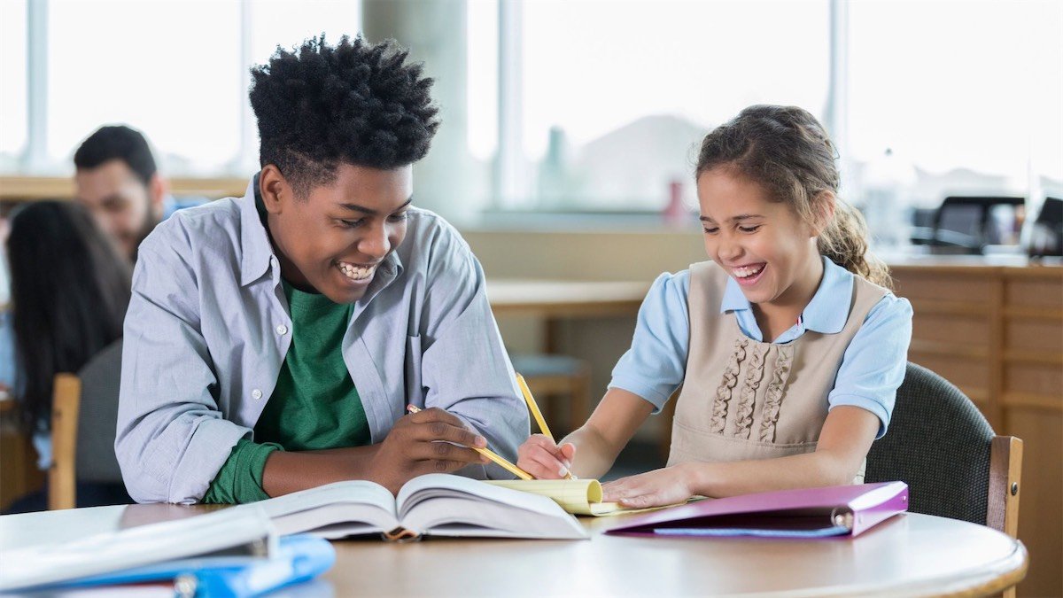 A young boy and girl sitting at a table in a classroom, smiling and working together with open books and notebooks.
