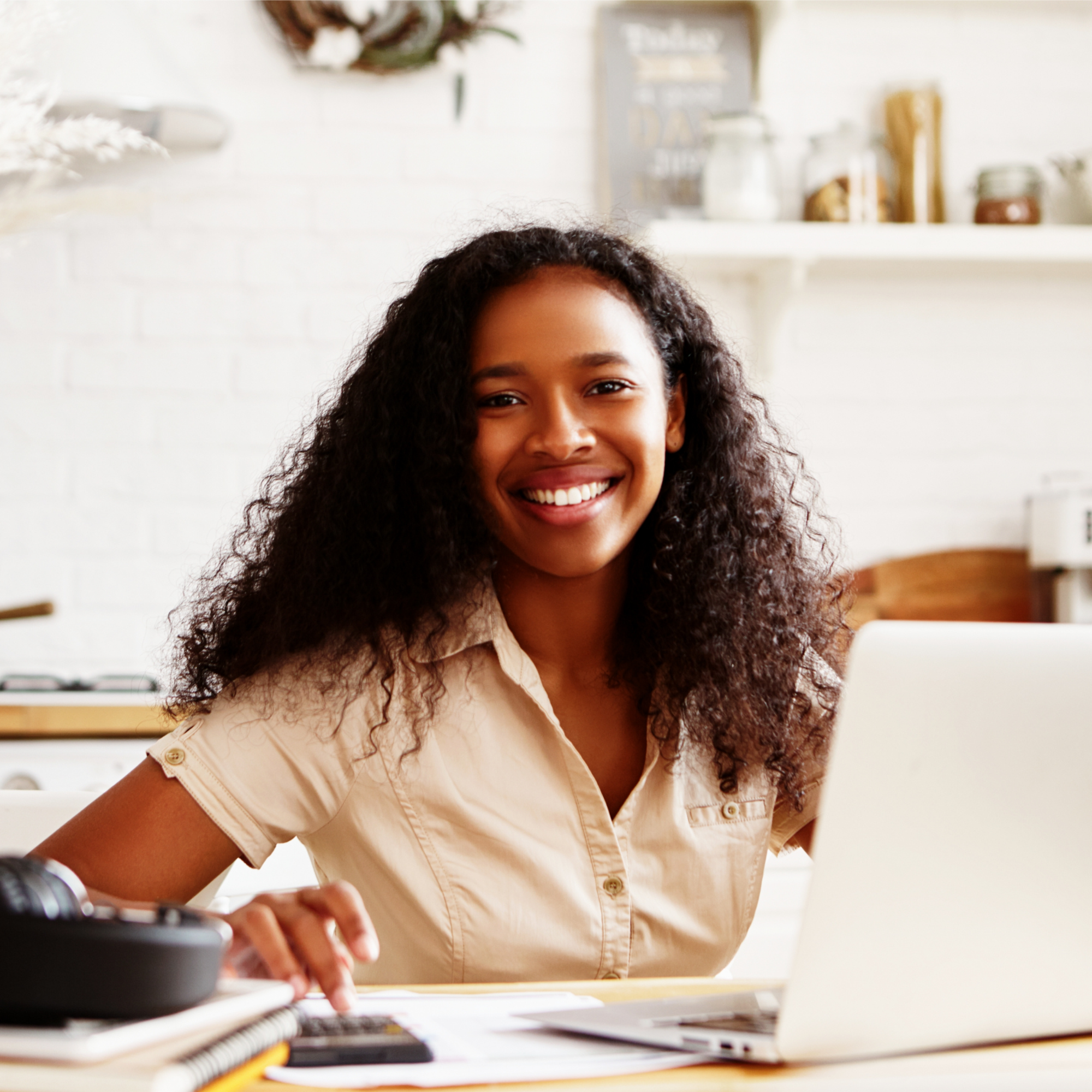 A woman with long curly hair smiling while looking at a laptop in a bright room with shelves and decorative items in the background.