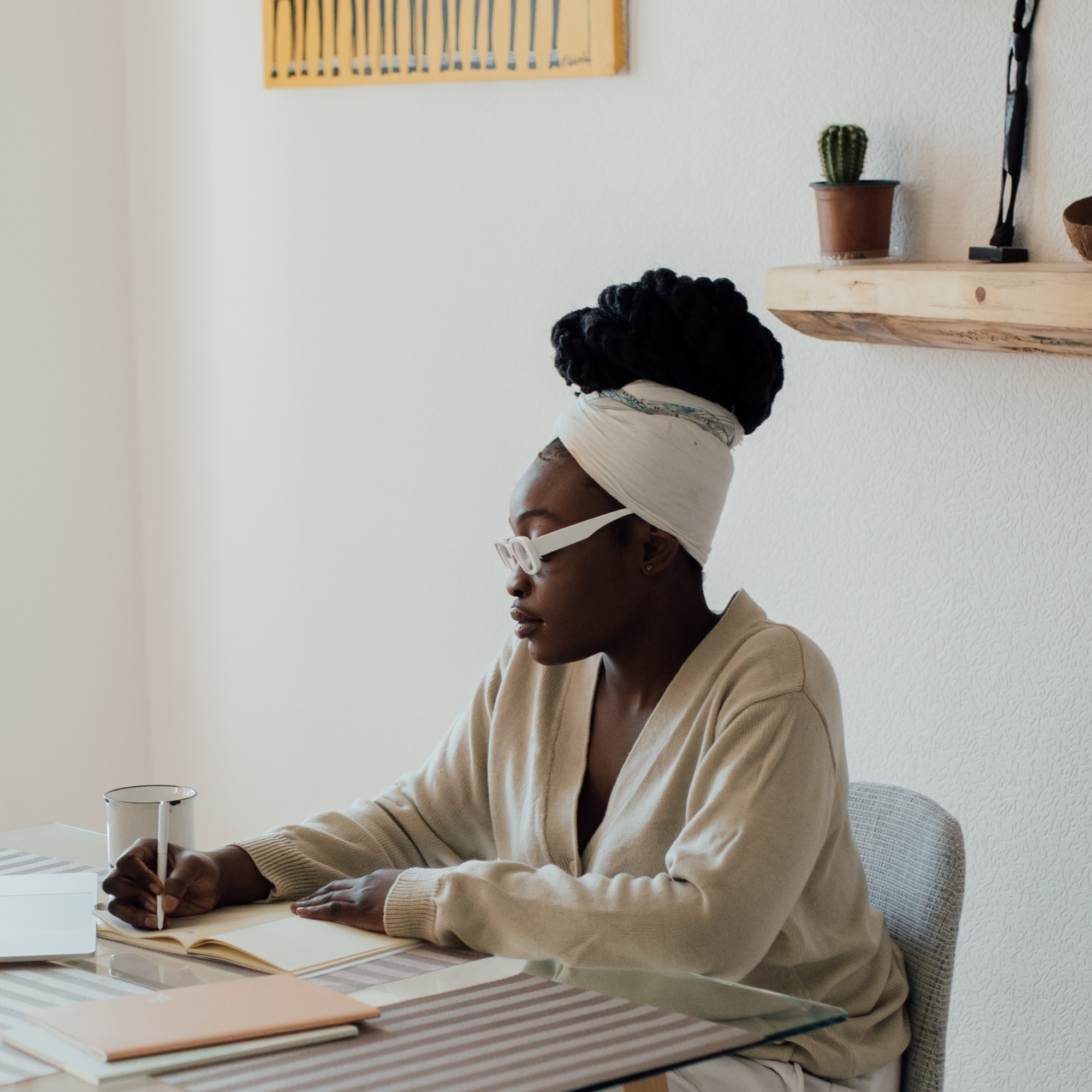 A woman with dark skin, glasses, and dreadlocks wearing a beige sweater sits at a table writing in a notebook with a pen. Her hair is wrapped in a beige headscarf. She is surrounded by books and a glass of water. The background features a white wall with a cactus in a pot on a wooden shelf and a piece of artwork.