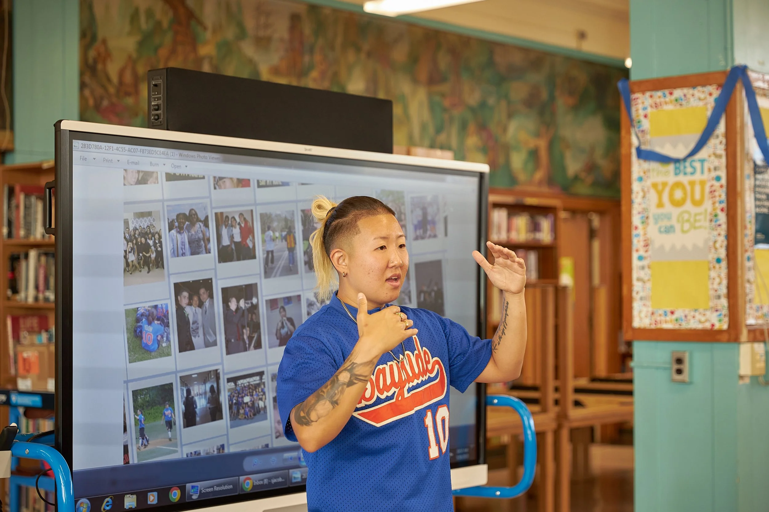 Jiggy Yoon speaking beside a projected collage while wearing a Bayside High School softball jersey