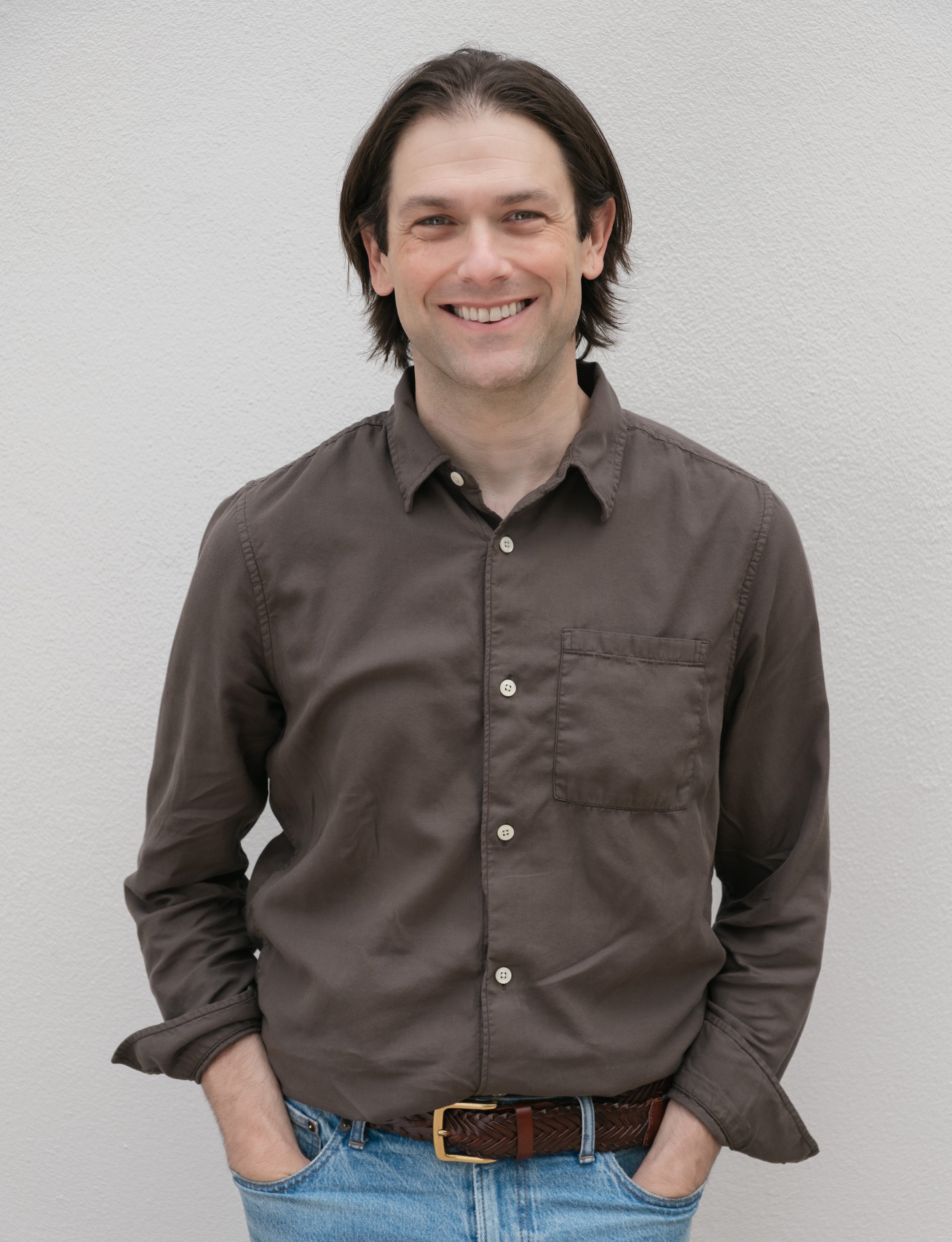 A man with dark hair wearing a brown button-up shirt and blue jeans, standing with his hands in his pockets against a plain white wall.