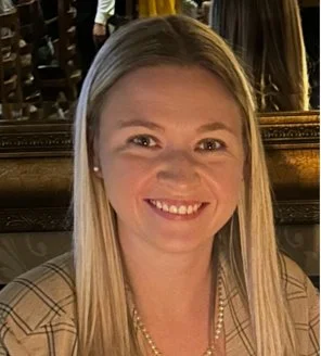 A smiling woman with long blonde hair, wearing a beige plaid blazer and a pearl necklace, sitting indoors with a mirror and other people in the background.
