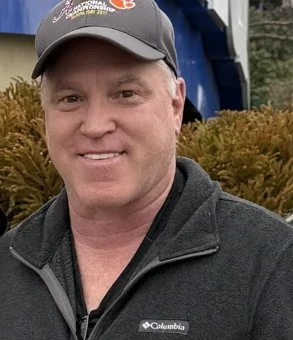 A smiling man wearing a baseball cap and a Columbia jacket outdoors, with bushes and a large blue machine or object in the background.