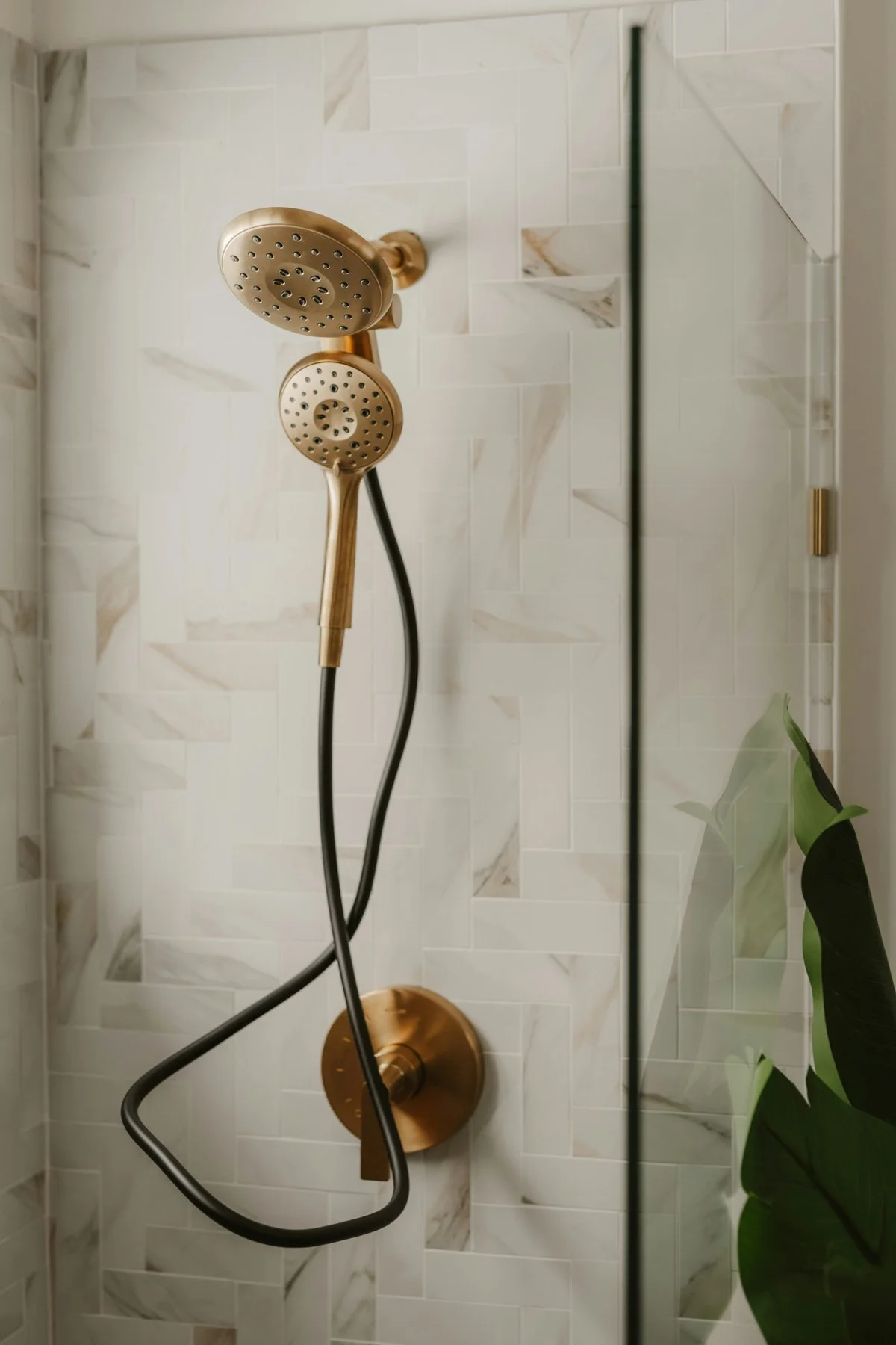 Brass-colored dual showerheads with a black hose on a tiled wall in a shower.