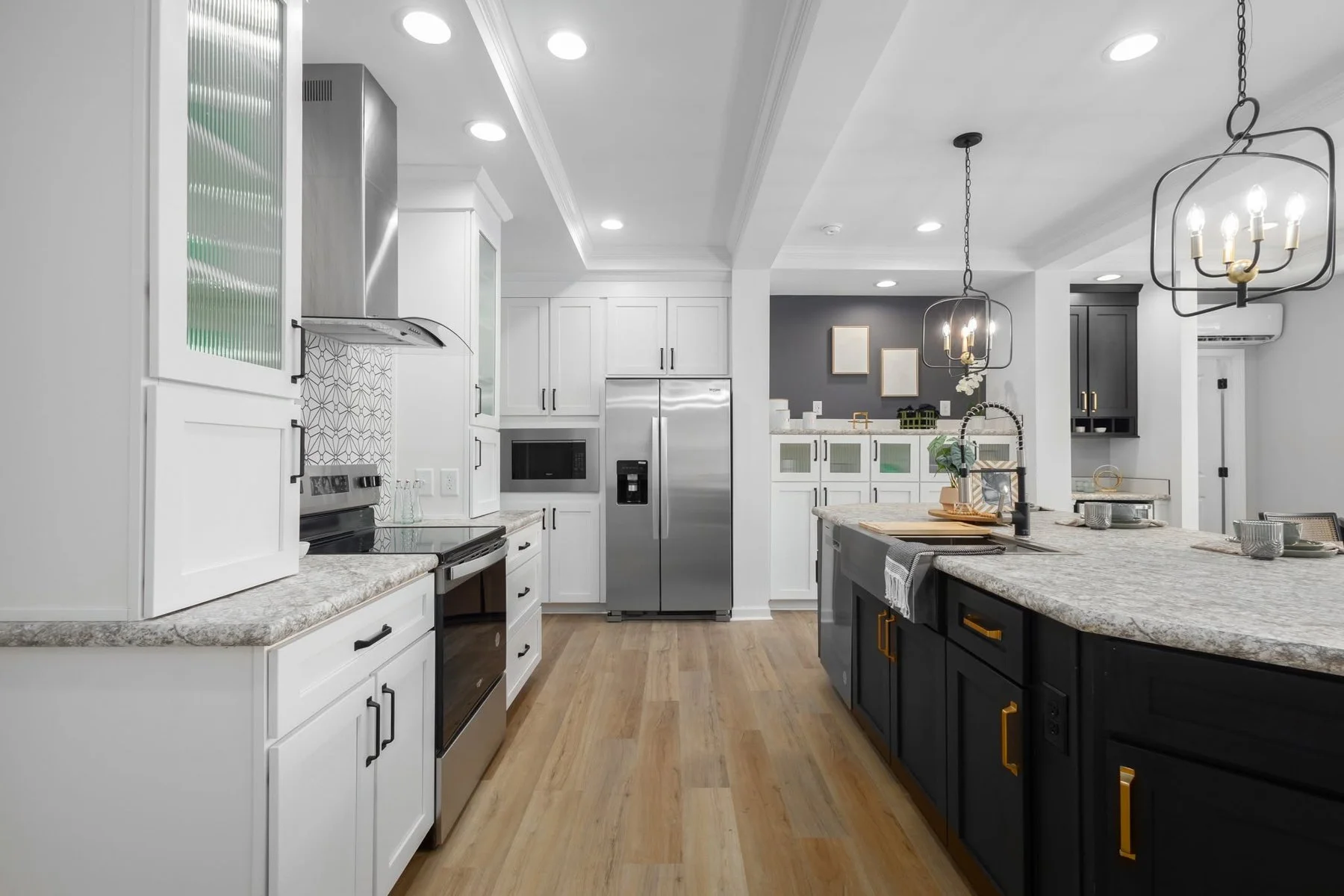 Modern kitchen with white and black cabinets, stainless steel refrigerator, marble countertops, wooden flooring, and black chandelier lighting.