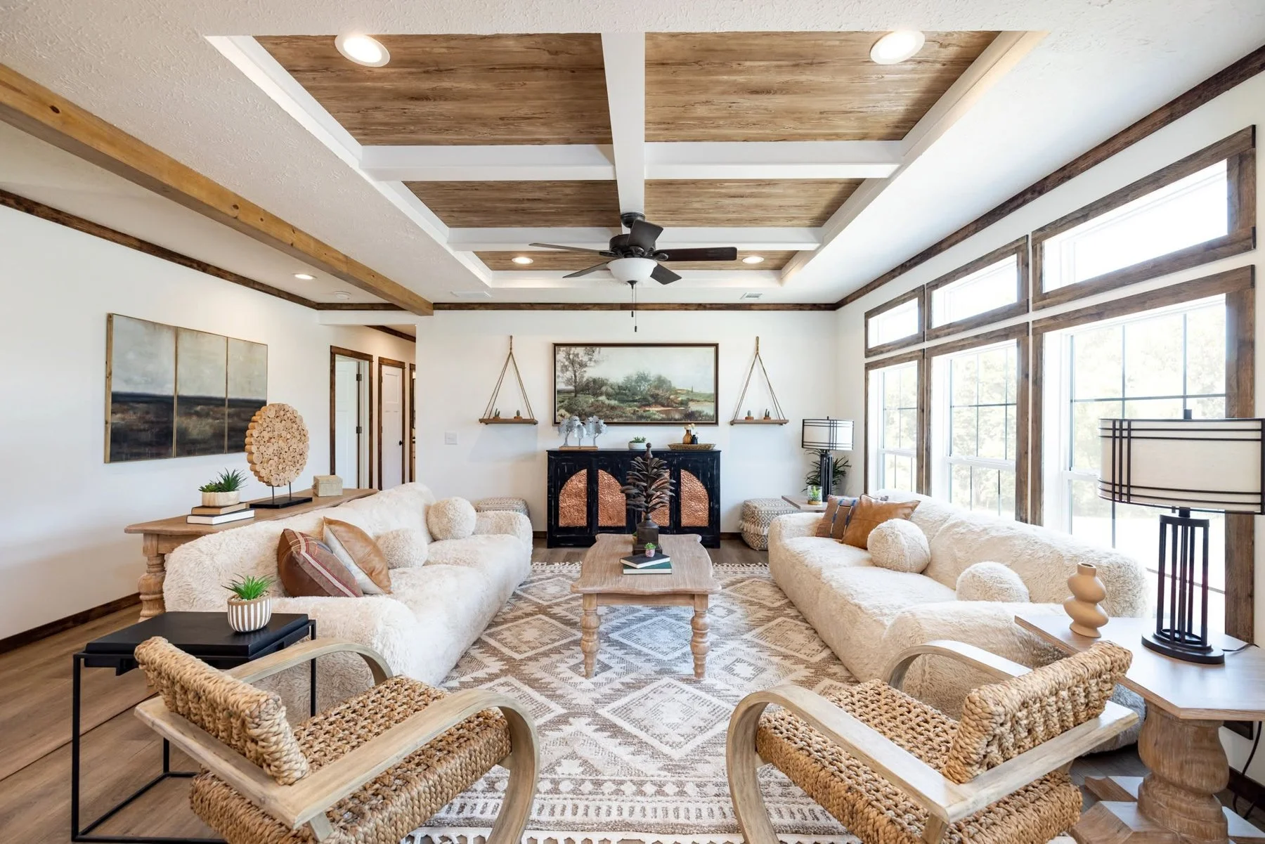 Living room with white plush sofas, wooden coffee table, and rattan armchairs. Large windows with wood trim, ceiling with wood panel accents, ceiling fan, artwork, and decor items like plants and lamps.