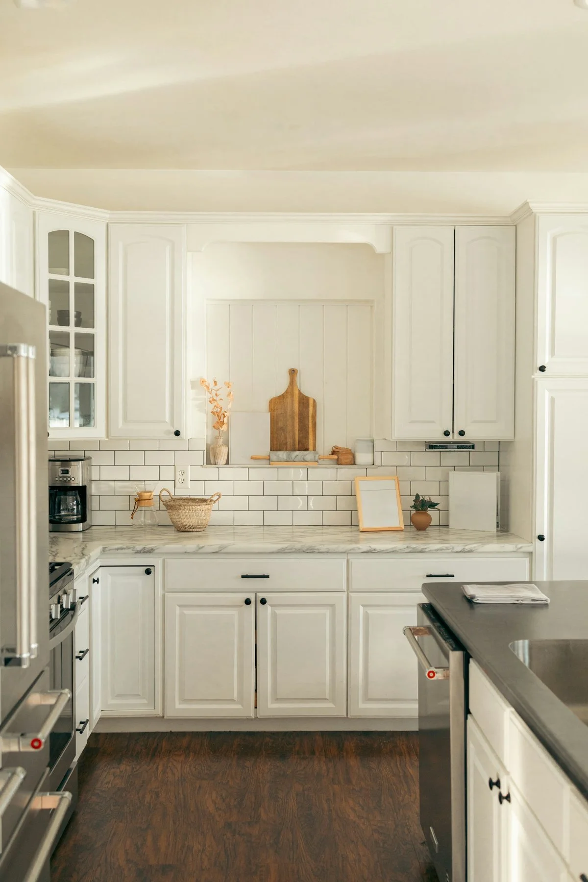 A white kitchen with upper and lower cabinets, marble countertop, white subway tile backsplash, and various kitchen items including a coffee maker, a basket, a wooden cutting board, and a small potted plant.