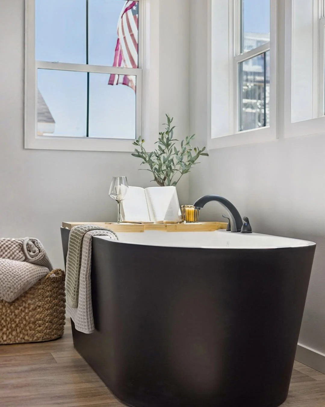 A modern black bathtub with a wooden tray holding an open book, wine glass, and a cup placed near a window with an outdoor view. A green plant is behind the tub, and a basket with folded towels is next to it.