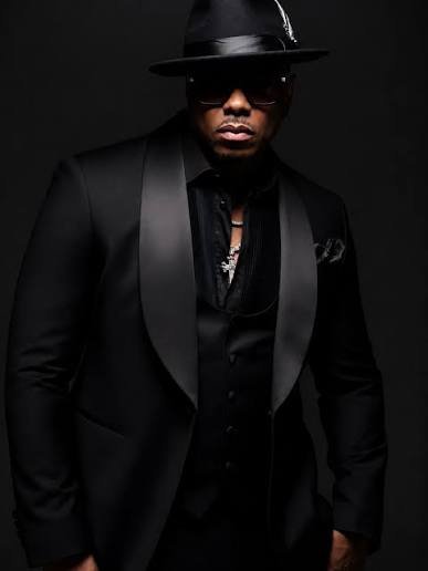 A man in a black suit, black shirt, and black hat with sunglasses, posing in front of a dark background.