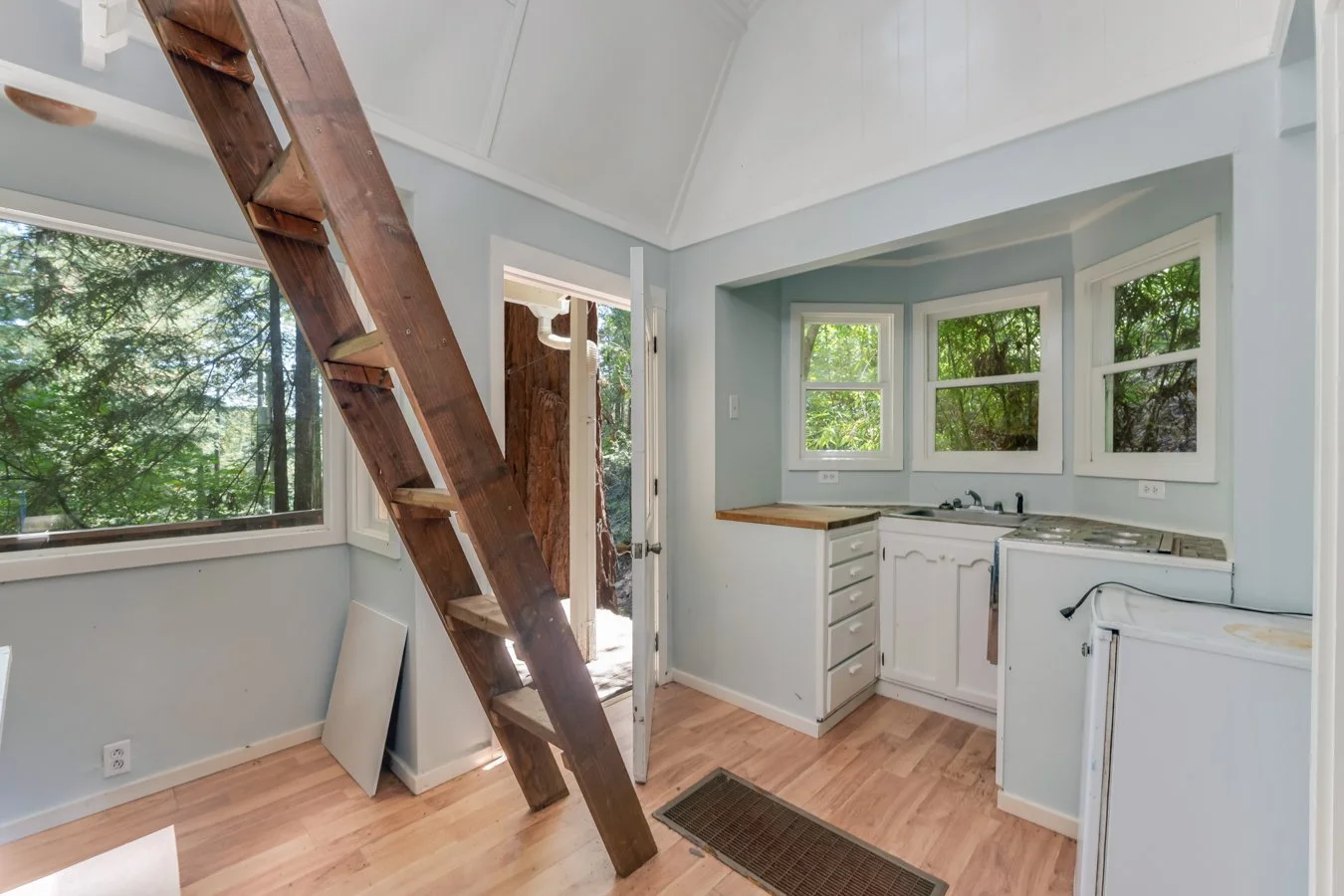 Small kitchen with light blue walls, white cabinets, three windows with greenery outside, wood countertop, and a slanted wooden staircase.