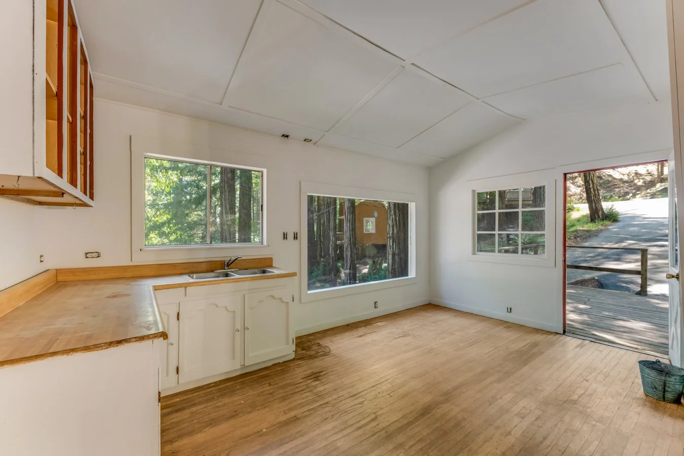 Empty kitchen with wooden floor and white walls, large windows showing trees outside, and an open door leading out to a deck.