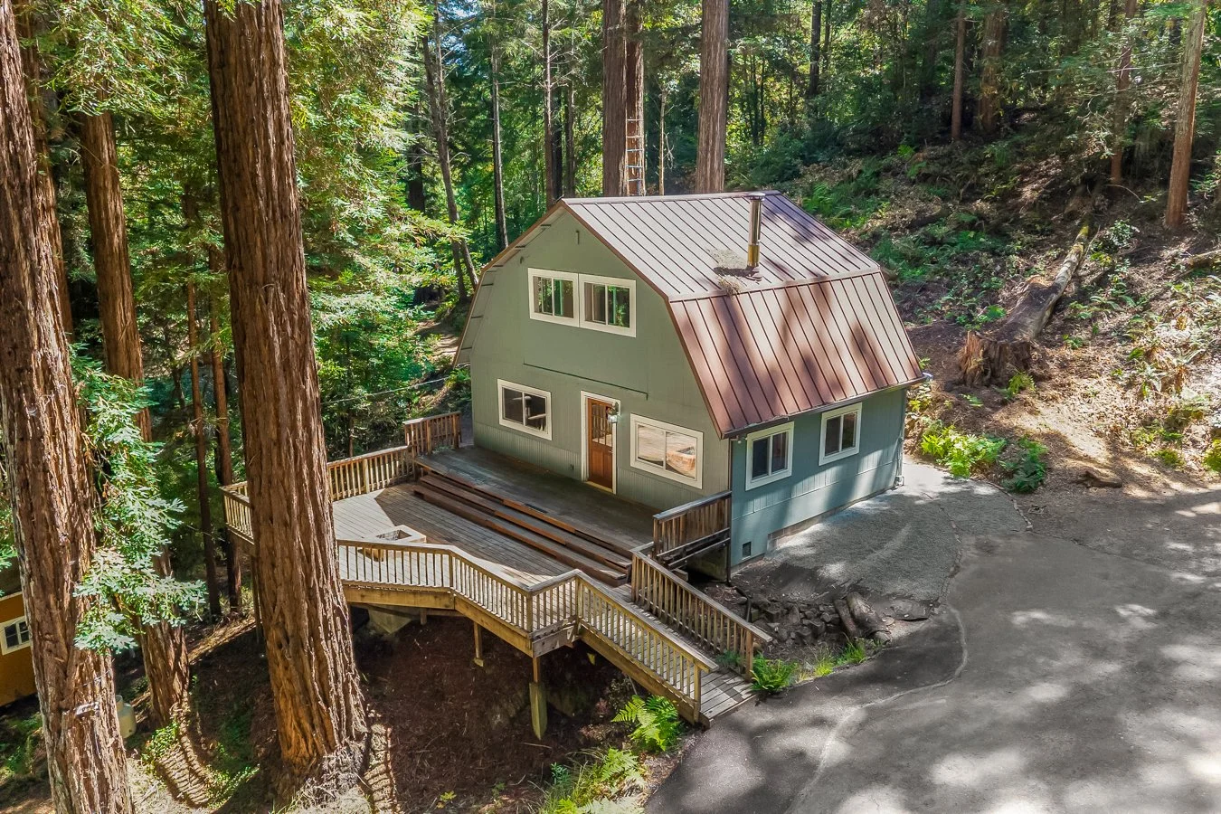 A two-story house with a metal roof, painted green, surrounded by tall trees in a forested area. It has a front porch with stairs and a wooden railing, and a sloped driveway leading to the house.